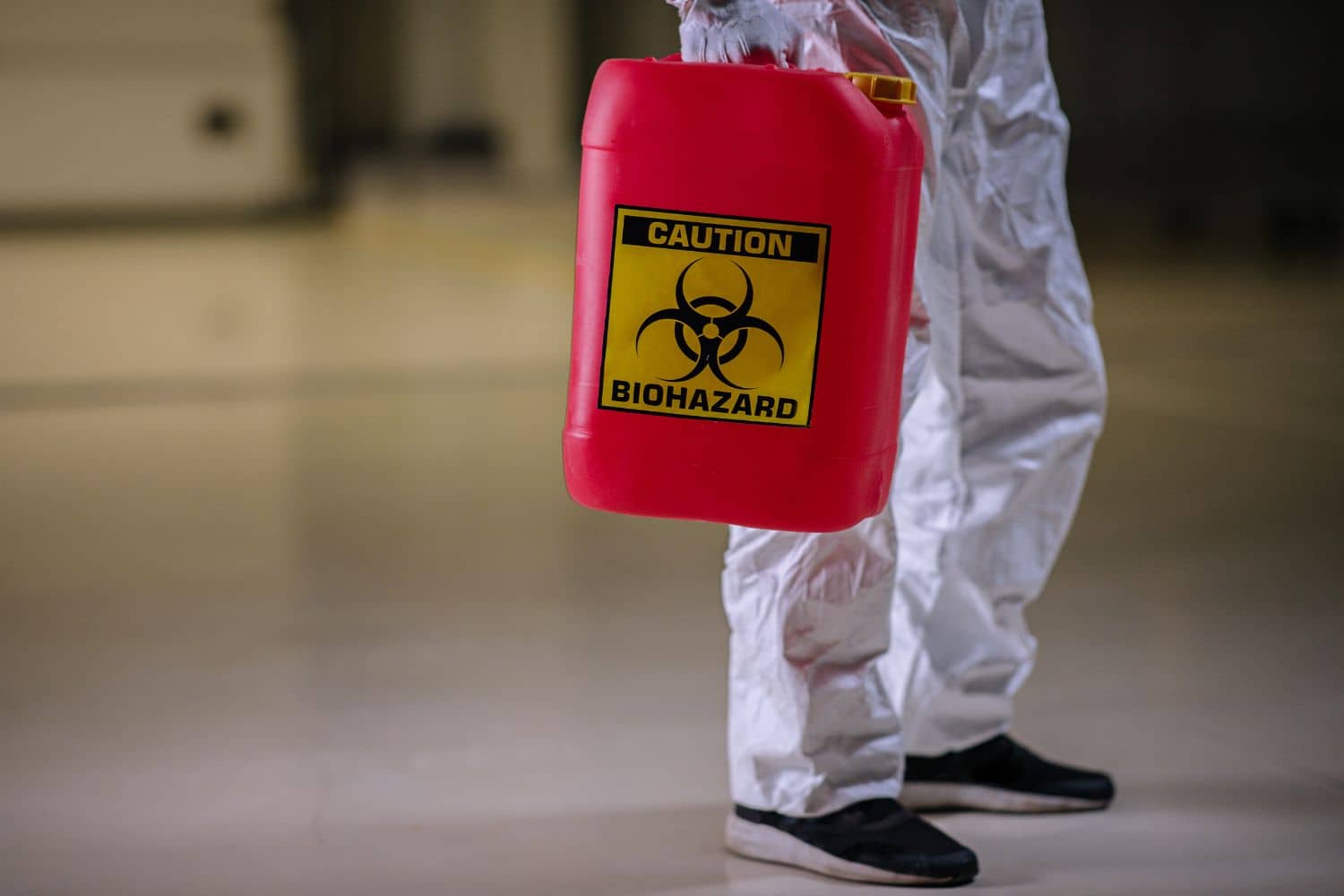 A person in a protective suit is holding a red container with a biohazard sign on it.