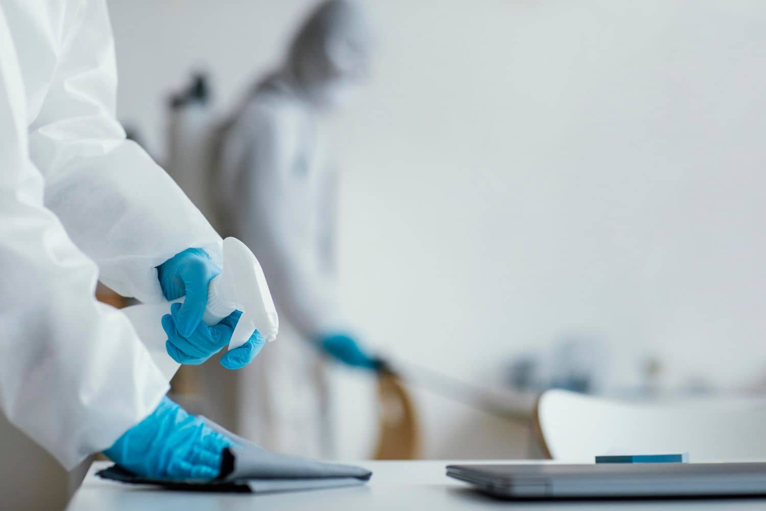 A person in a protective suit is cleaning a desk with a spray bottle.