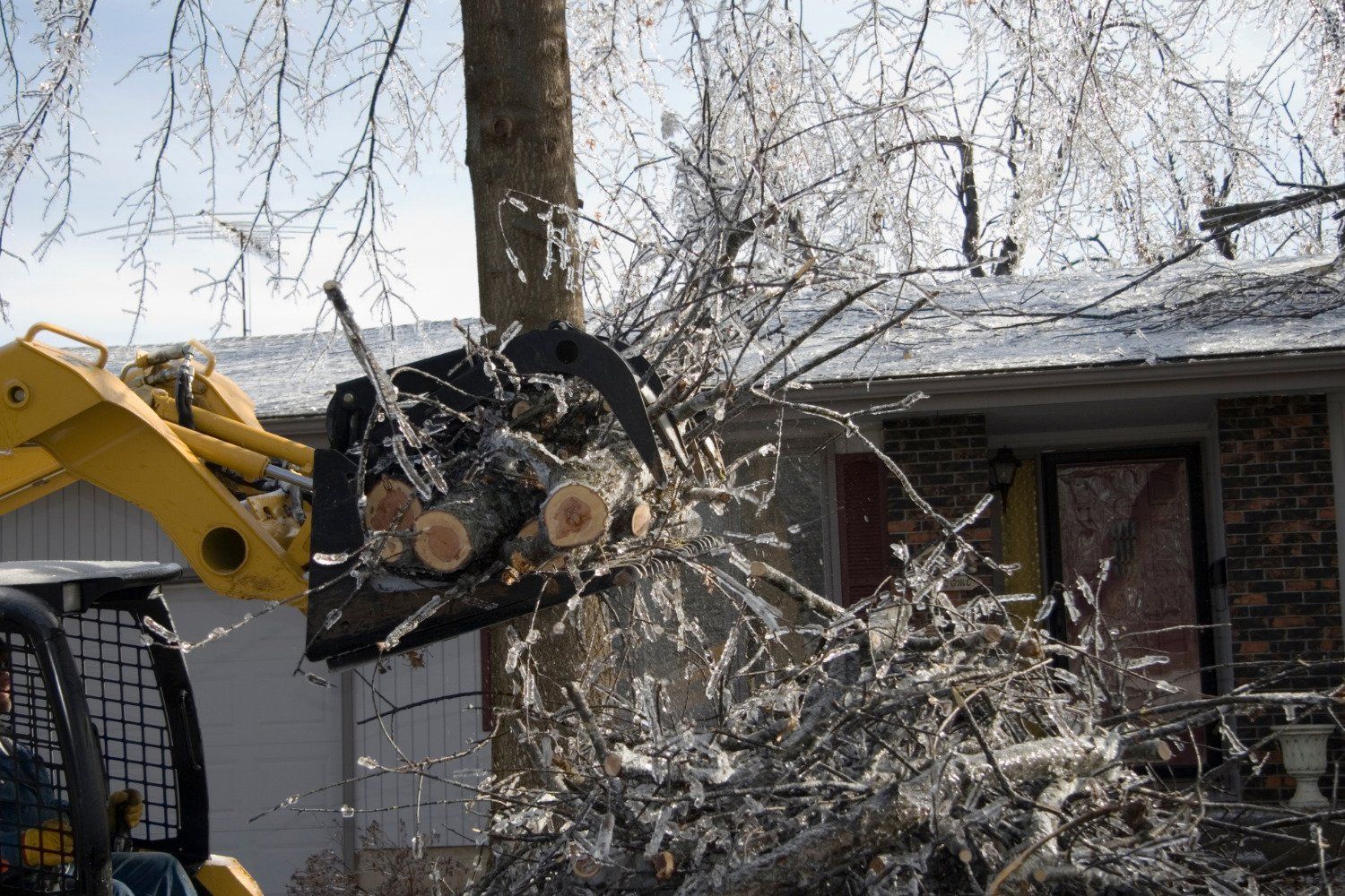 A yellow tractor is cutting a tree in front of a house.