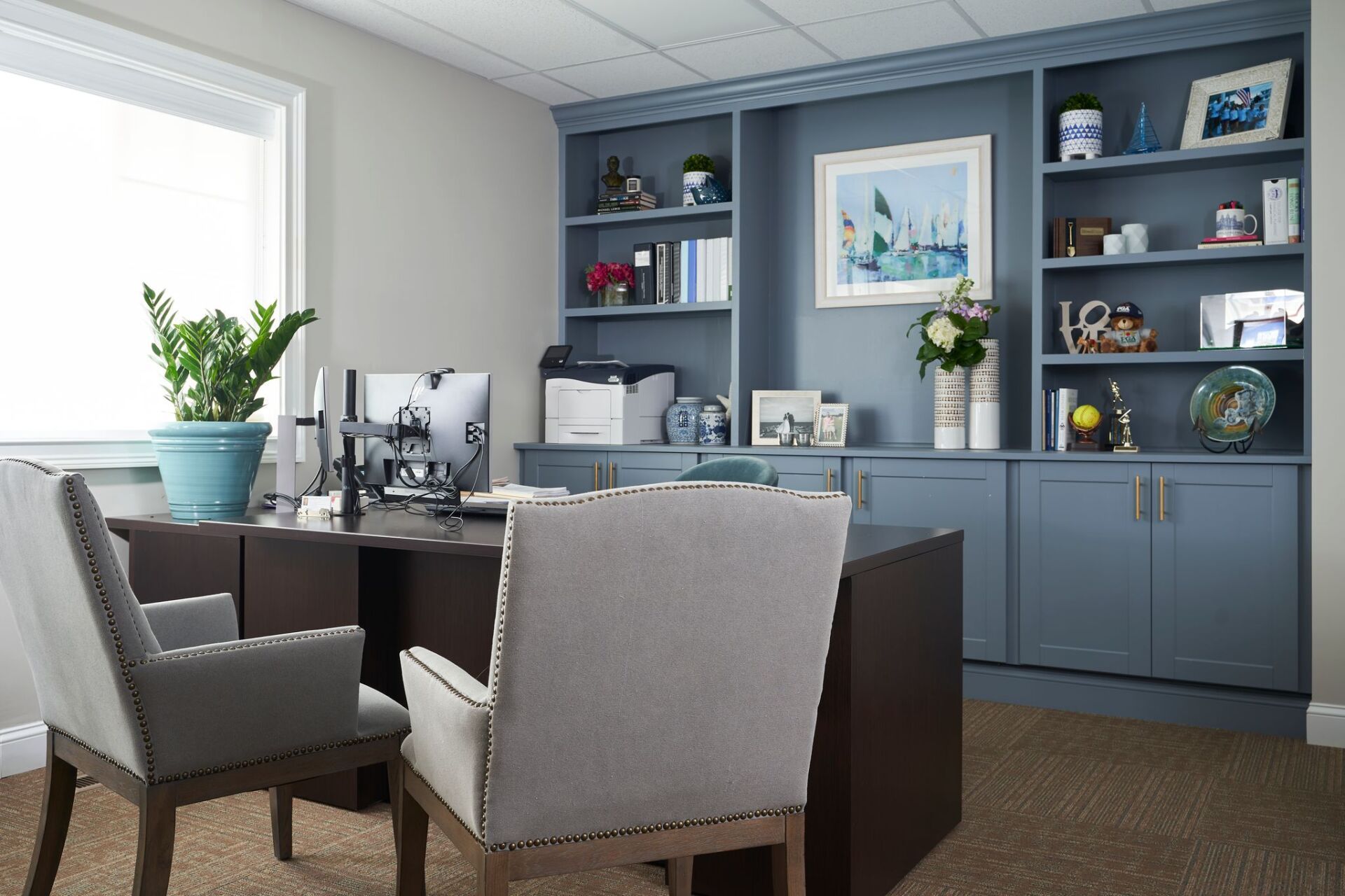 Office with dark desk, grey chairs, built-in blue shelves filled with decor, and a window.