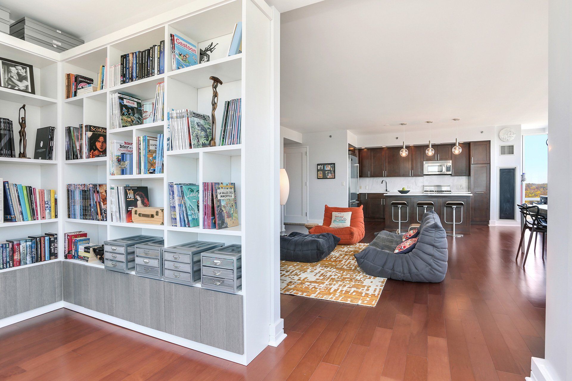 White bookshelf with books, opening into a living space with bean bag chairs and a kitchen.