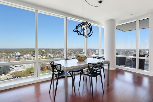 Dining room with large windows overlooking a city, featuring a table, chairs, and modern light fixture.
