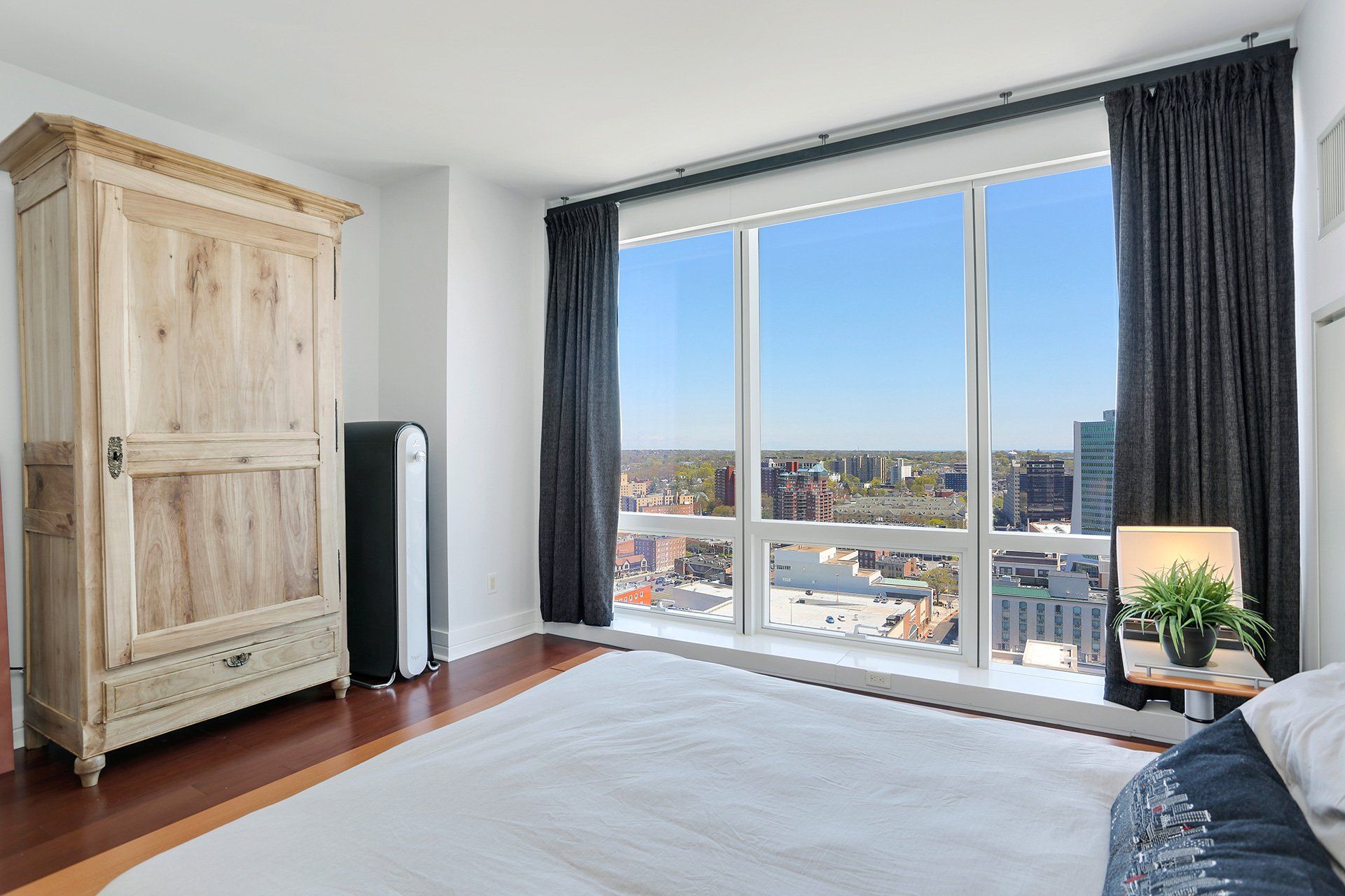 Bedroom with tall windows, wooden armoire, white rug, and city view.
