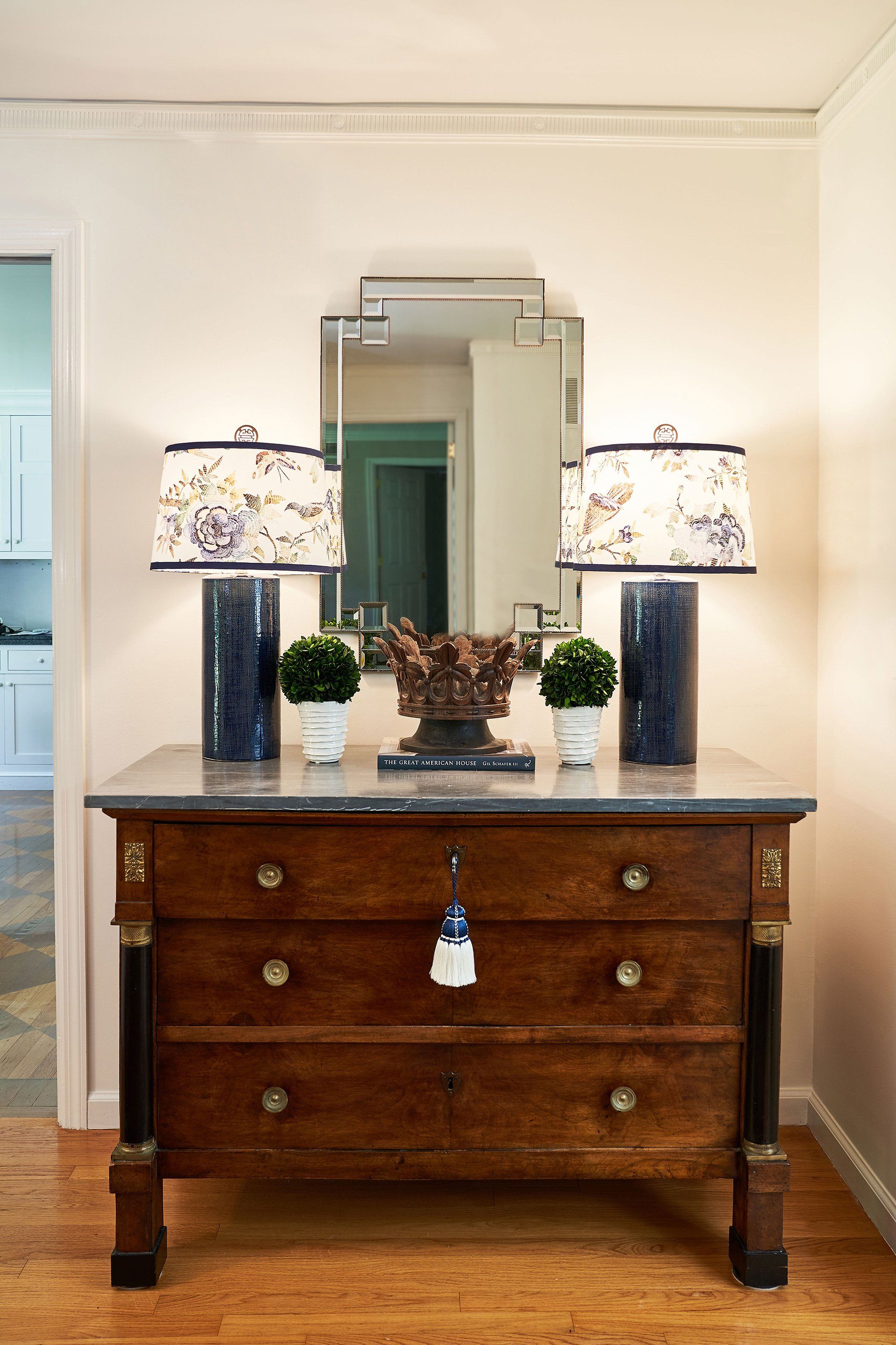 A wooden dresser with a marble top, flanked by lamps, a mirror, and decorative plants.