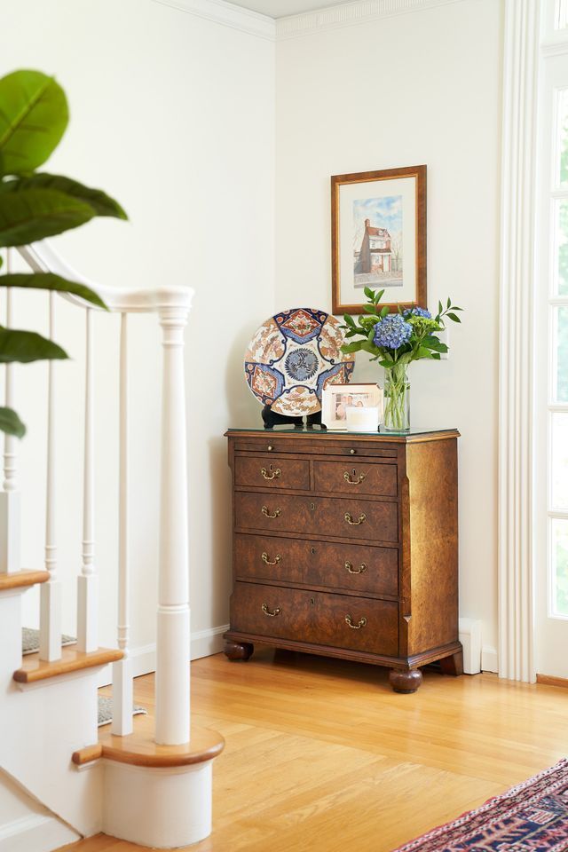 Wooden chest of drawers in a corner, with a decorative plate and flowers. Near a staircase and window.