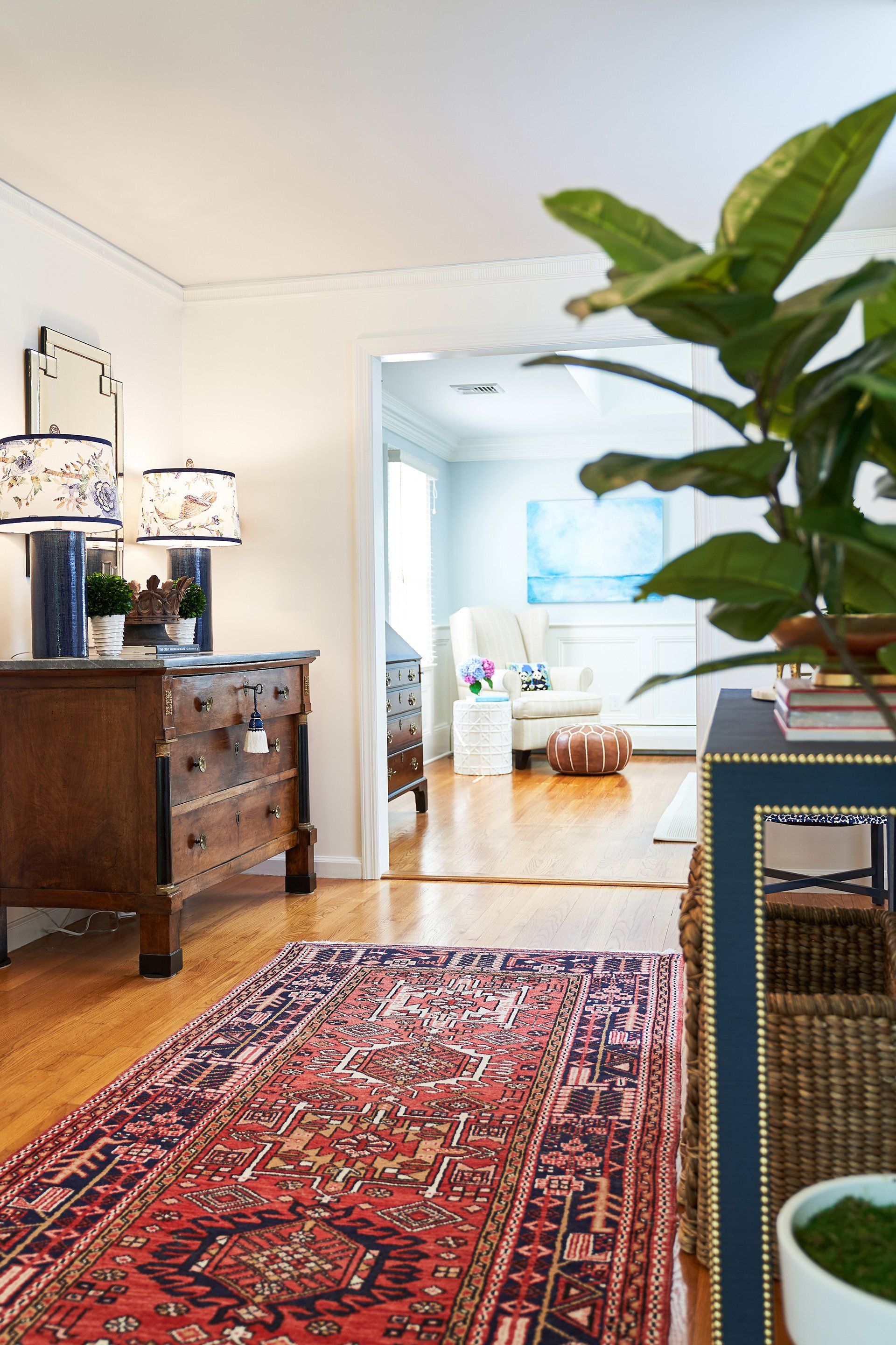 Entryway with antique dresser, oriental rug, blue lamps, and a glimpse of a light-filled room.