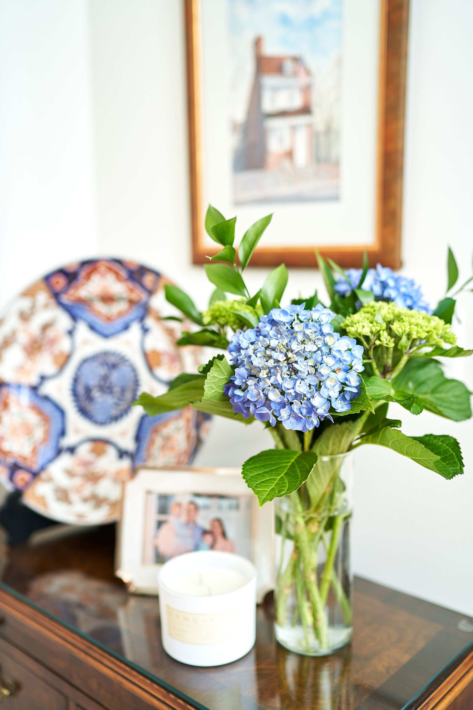 Hydrangeas in a glass vase on a wooden table with a plate, framed photo, and candle, under a framed picture.