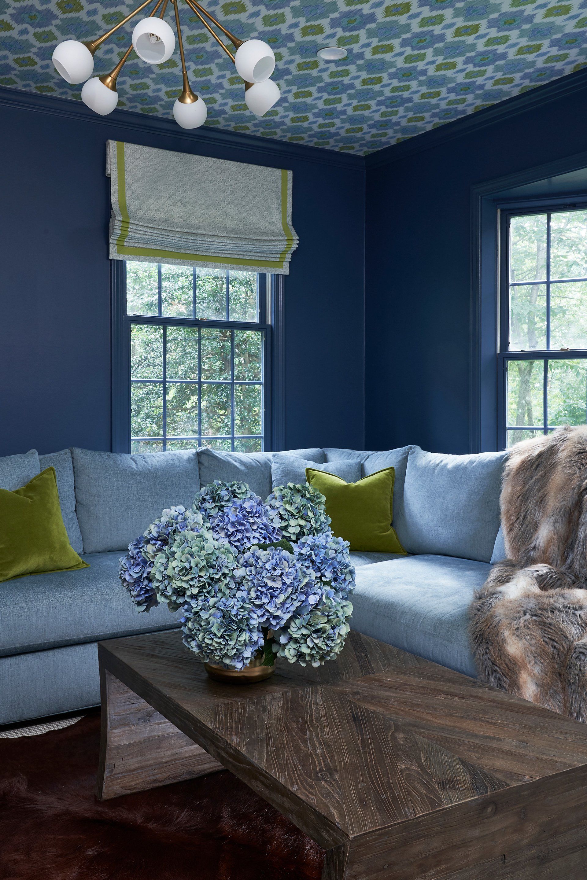 Blue-walled living room with sectional sofa, floral arrangement, and patterned ceiling. Windows look out onto greenery.