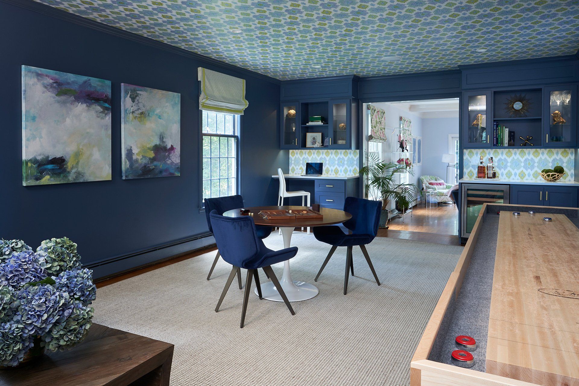Blue room with round table, chairs, art, and shuffleboard. Patterned ceiling and rug.