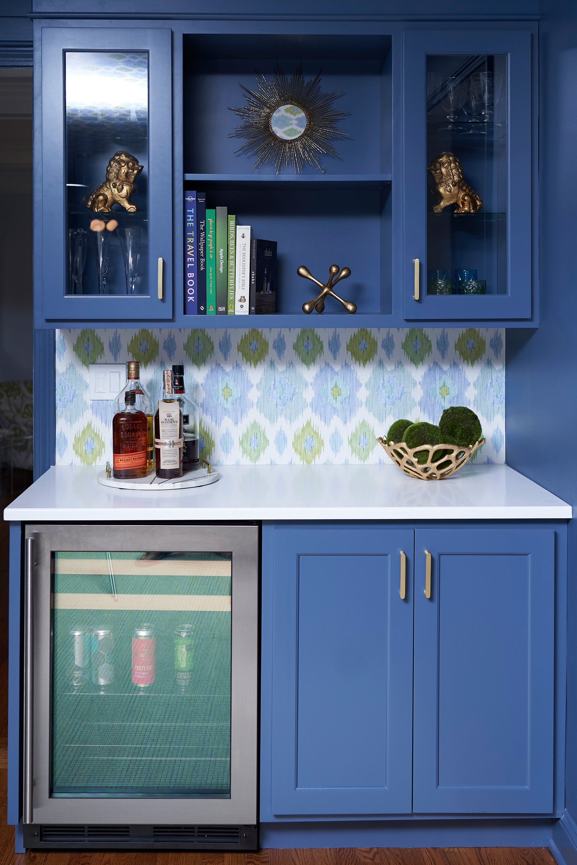 Blue home bar with upper cabinets, countertop, and wine cooler. Backsplash and decorative items.