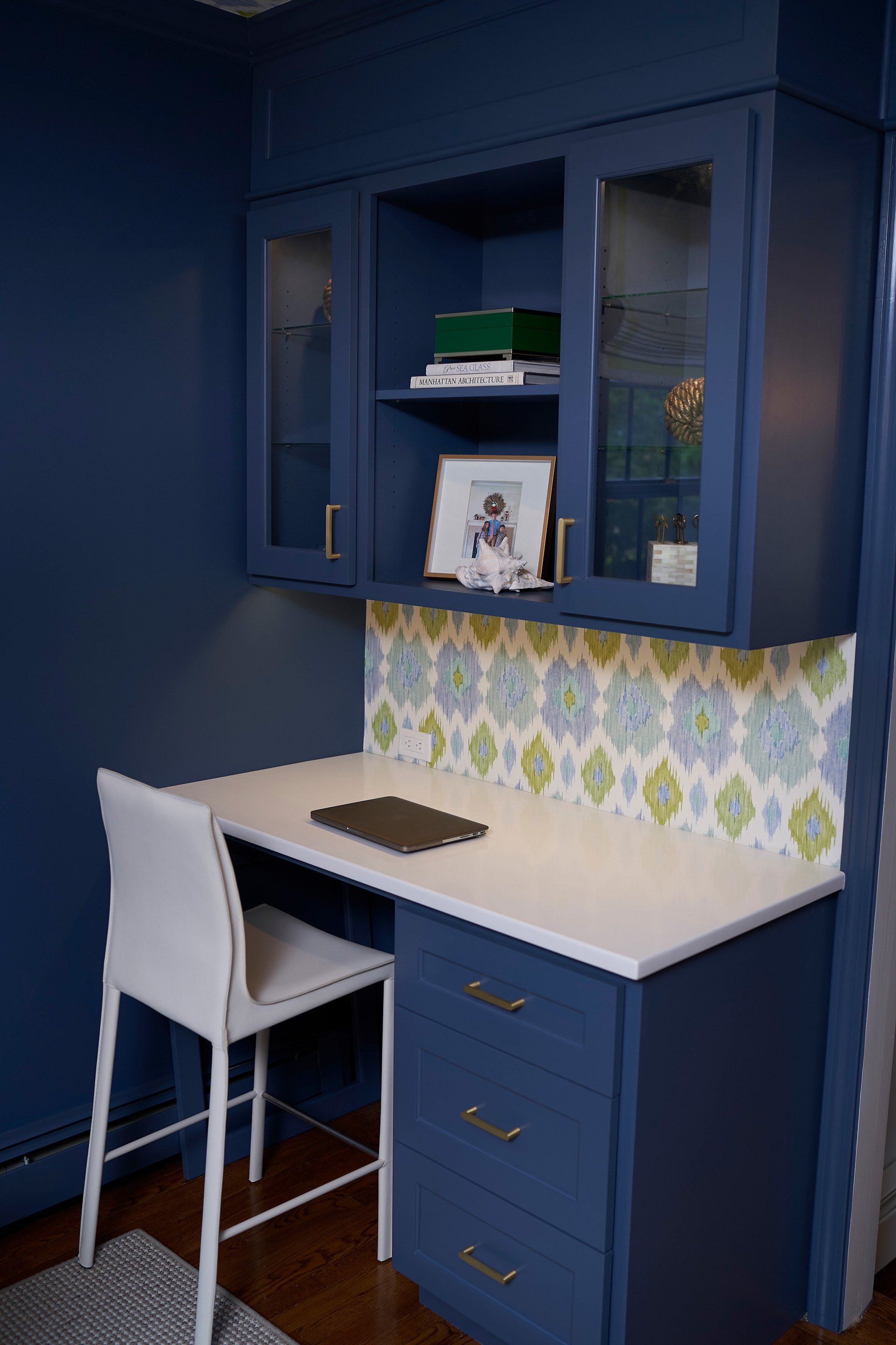 Blue home office with a white desk, chair, and cabinets with gold handles. Wallpaper backsplash.
