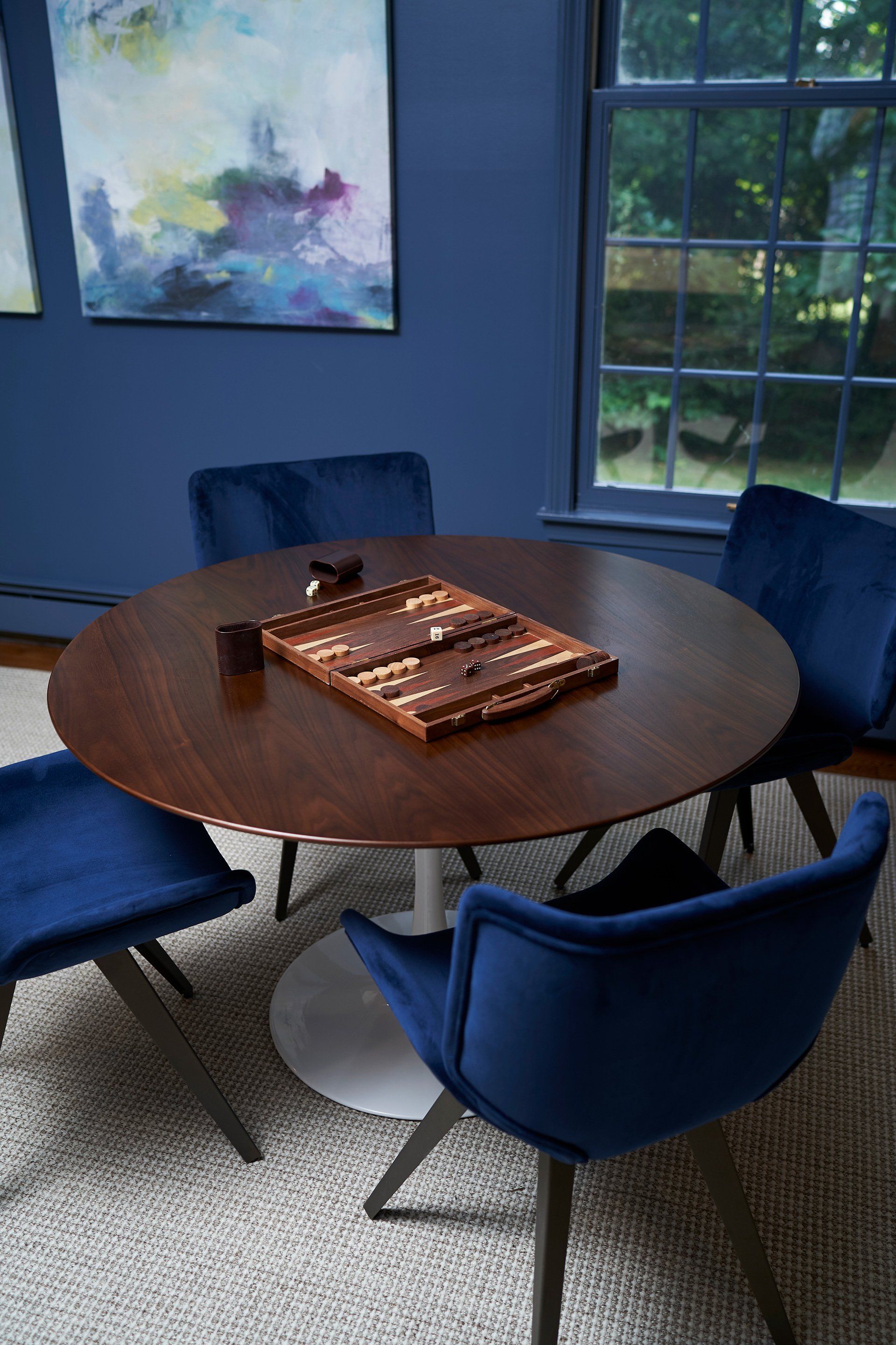 Round wood table with backgammon set, surrounded by four blue velvet chairs in a room with blue walls and a window.