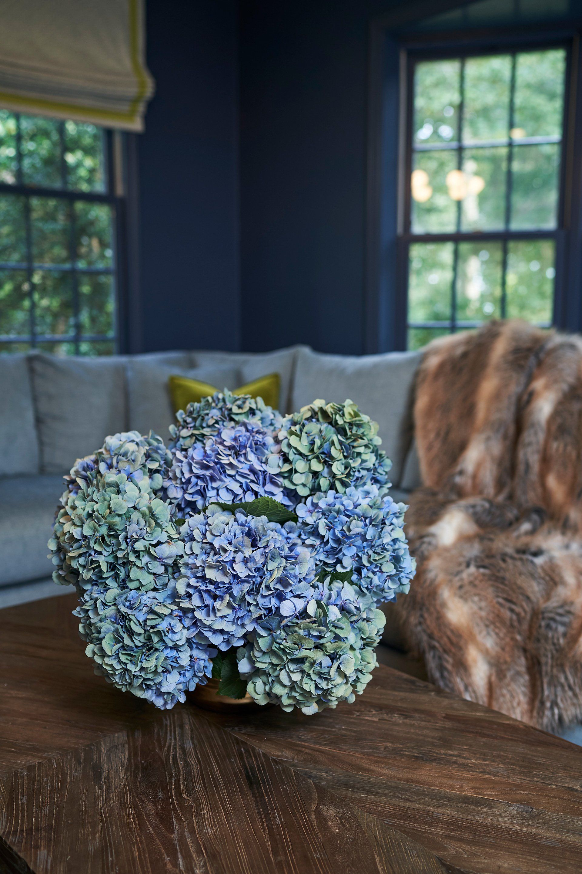 Blue and green flower arrangement on a dark wood coffee table in a navy blue room with grey couch and faux fur throw.