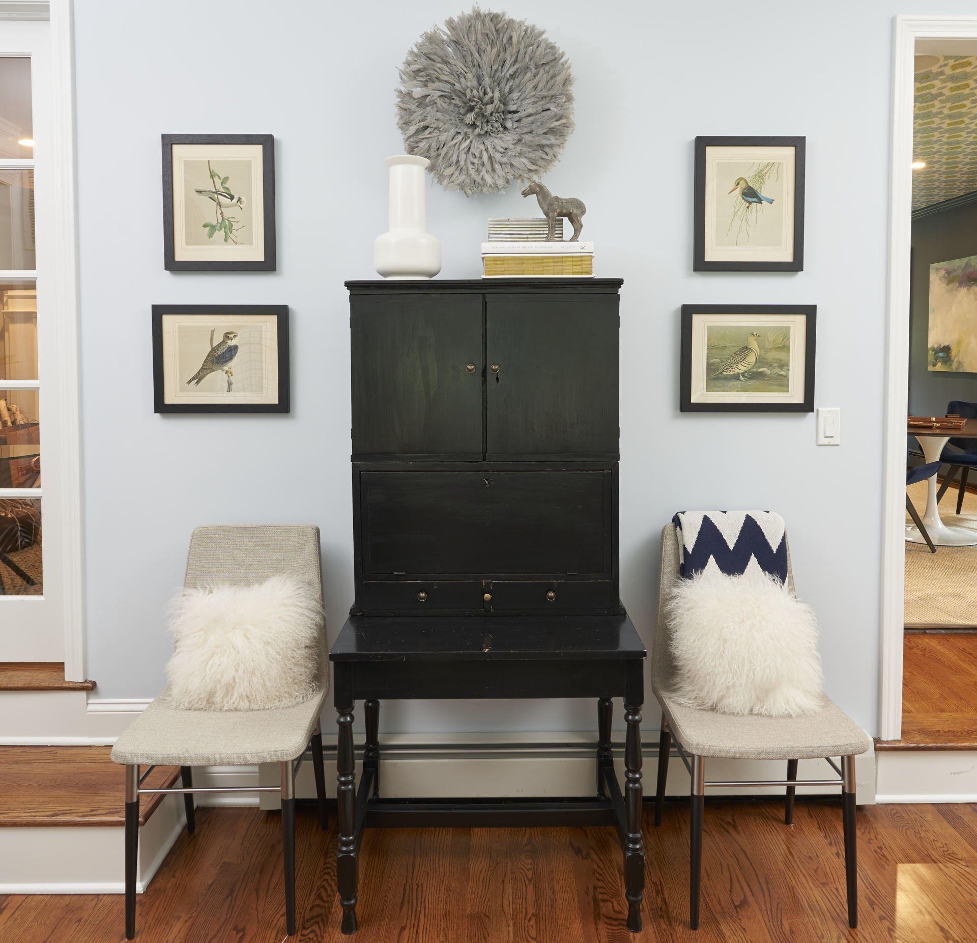 Black cabinet with framed art on a light blue wall, flanked by chairs with fluffy pillows.