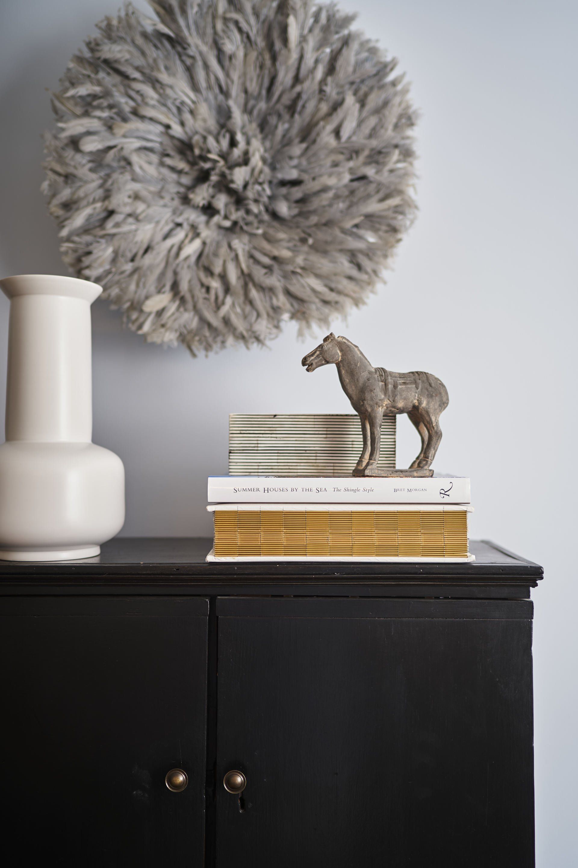 Black cabinet with decor: vase, horse statue on books, and gray feather wall art.