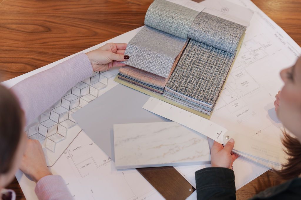 Two people reviewing design samples on a table with blueprints, fabric swatches, and a tile sample.