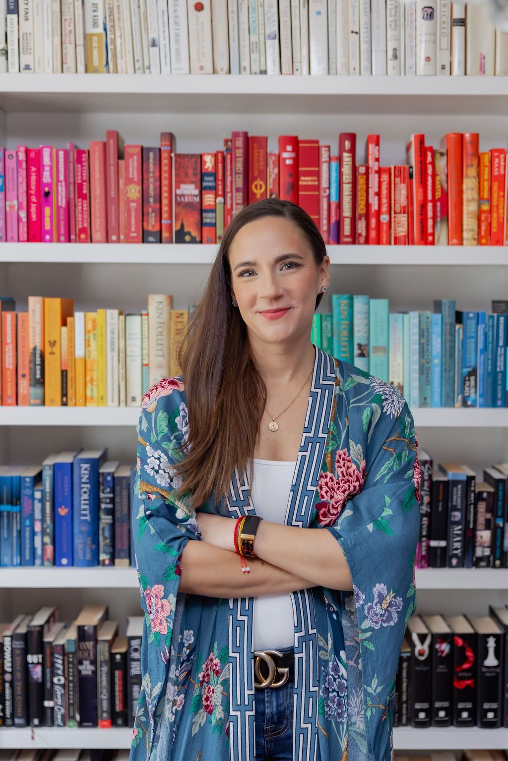 Woman with arms crossed in front of a colorful bookshelf. She wears a floral jacket and smiles.