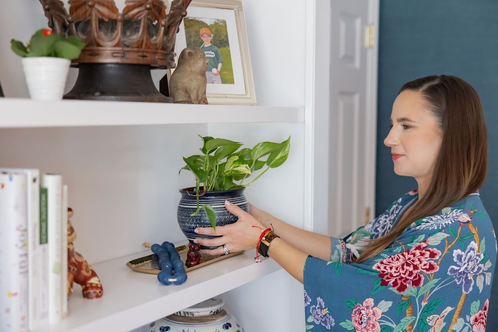 Woman arranging a potted plant on a white shelf. She wears a floral robe.