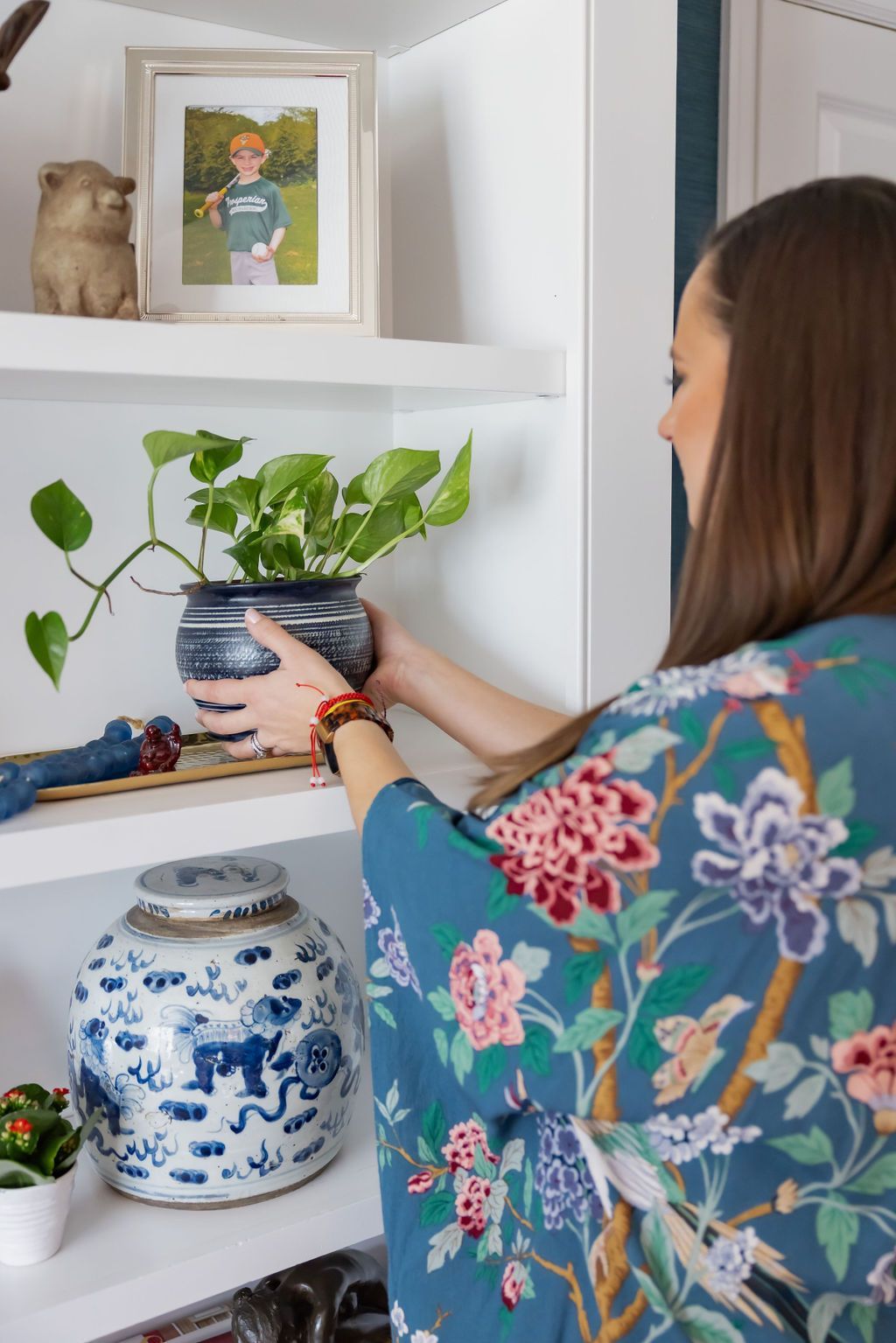 Woman arranging potted plant on a white shelf, wearing a blue floral robe.