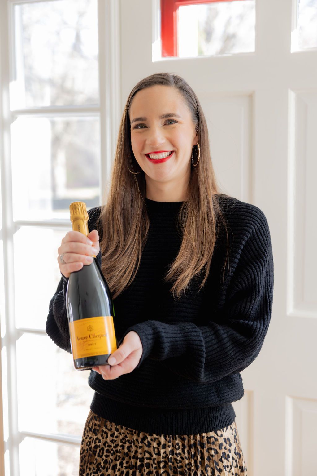Woman holding a champagne bottle, smiling. She wears a black sweater and leopard-print skirt, in a brightly lit room.