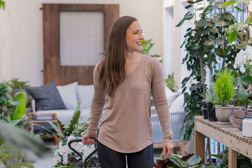 Woman smiles, walking through a plant-filled greenhouse; wooden furniture, shelves, and a gray-brown long-sleeved shirt.