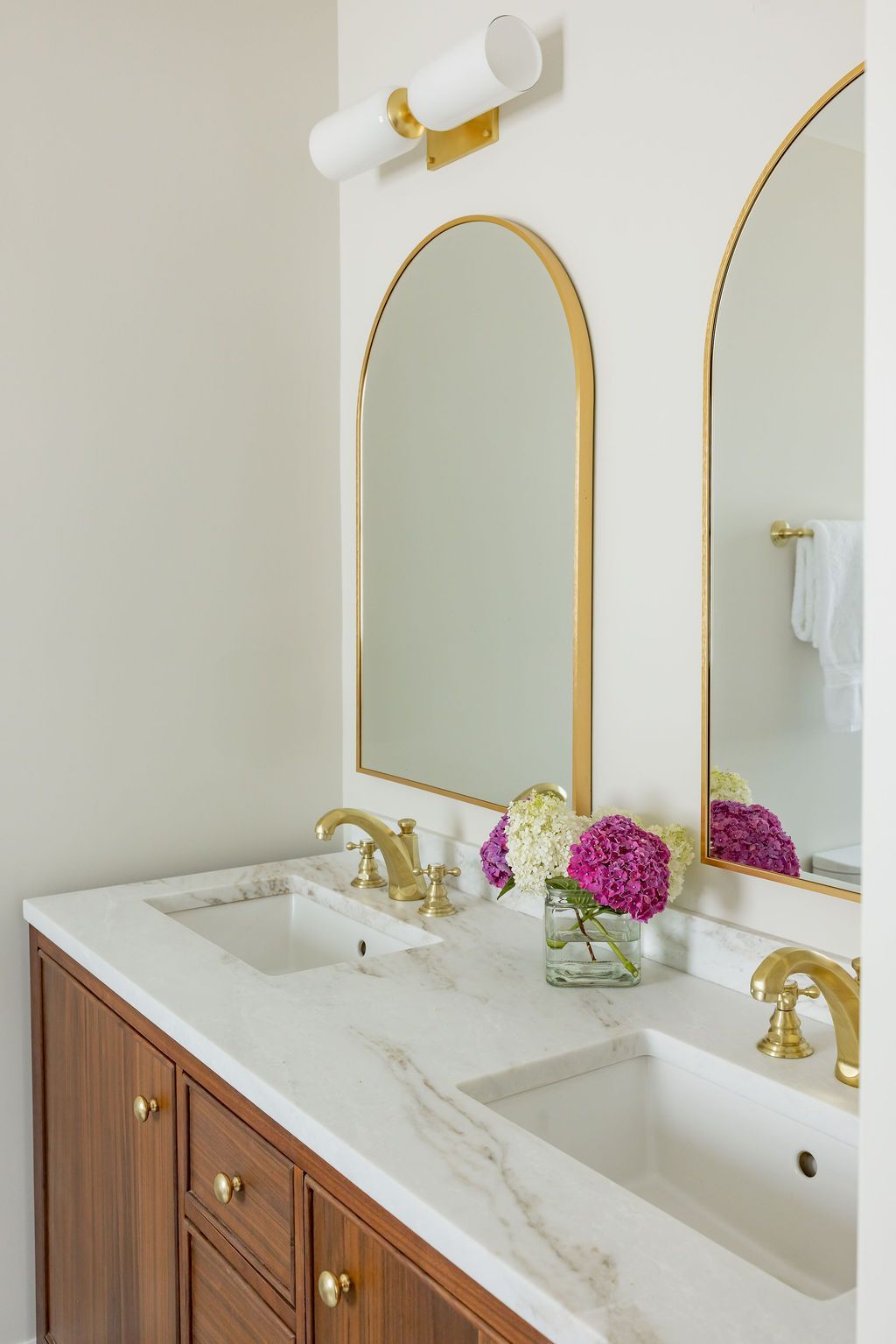 Bathroom with two sinks, arch mirrors with gold frames, and gold fixtures. Wooden cabinets, white countertop.