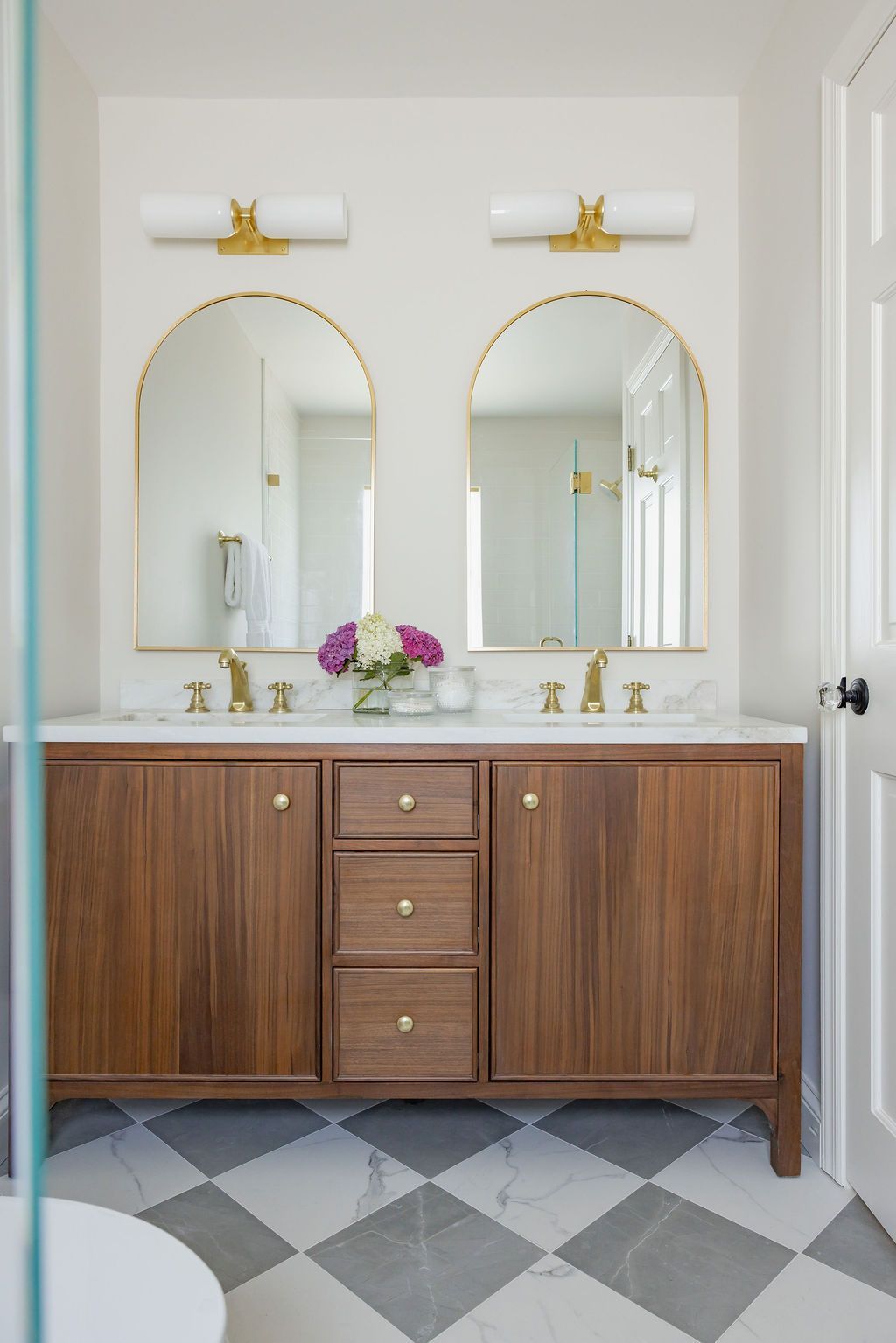Bathroom with dual sinks, arched mirrors, and wood vanity with drawers. Checkered floor and white walls.