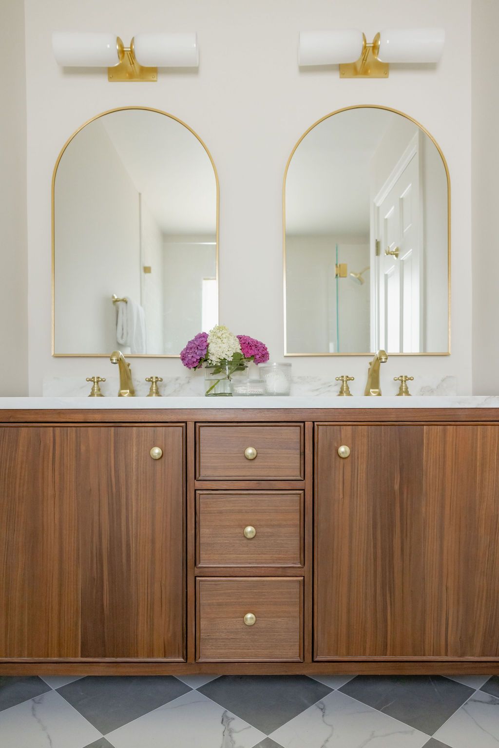 Bathroom with two arched mirrors, gold fixtures, wooden vanity, and a flower arrangement.