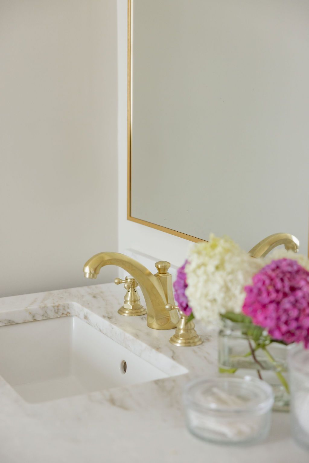 Gold faucet on a marble countertop in a bathroom, with a mirror and flowers.