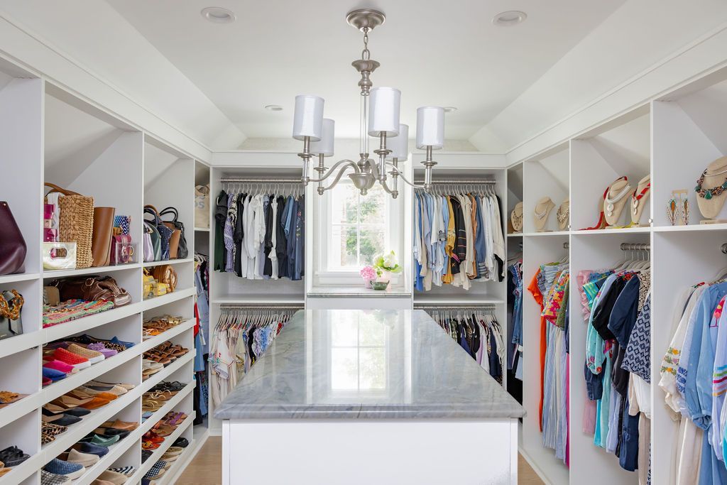 Walk-in closet with shelves of shoes and handbags, hanging clothes, and a central countertop, chandelier above.