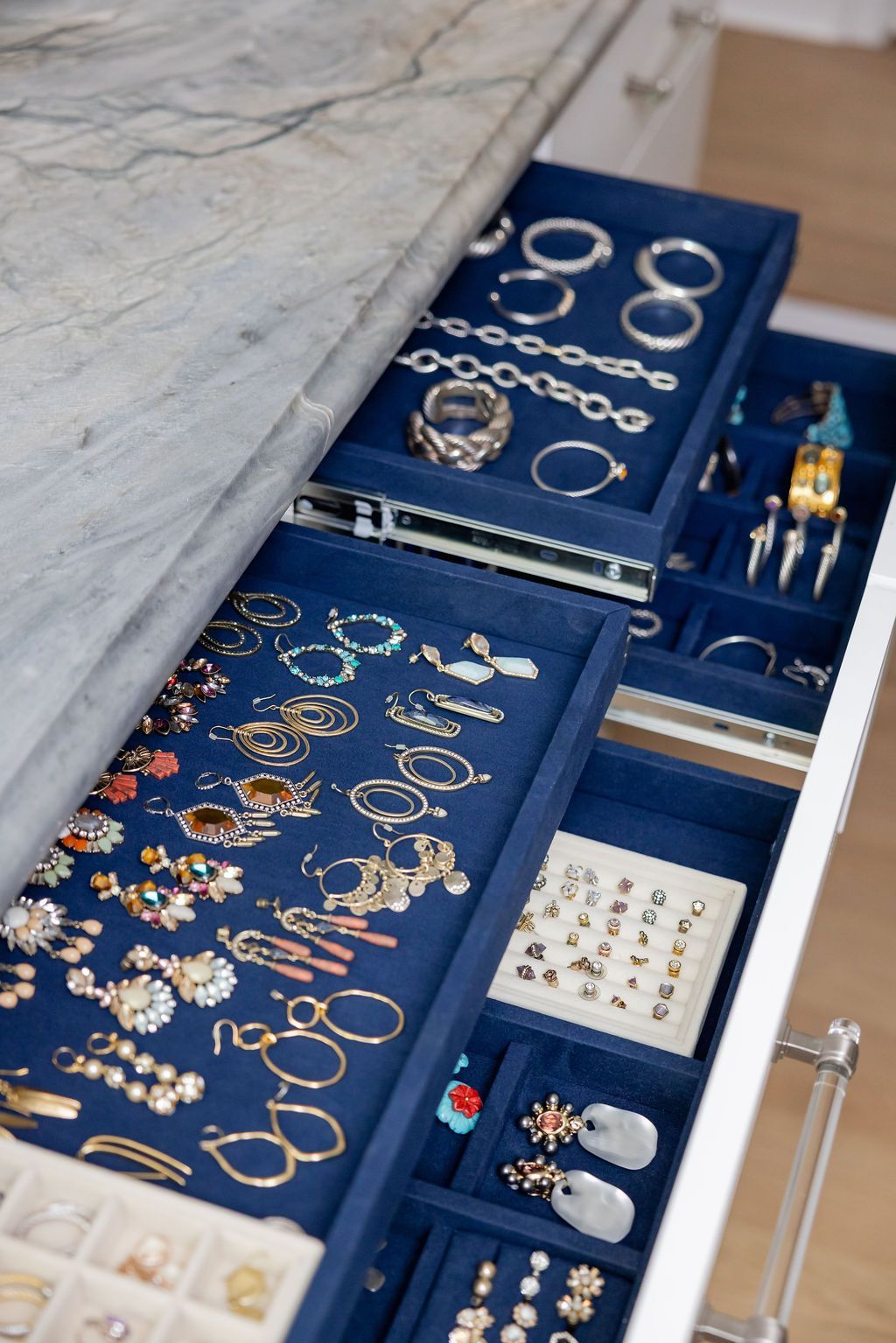 Jewelry drawer with blue velvet trays, displaying rings, bracelets, earrings, and necklaces, set on a white counter.
