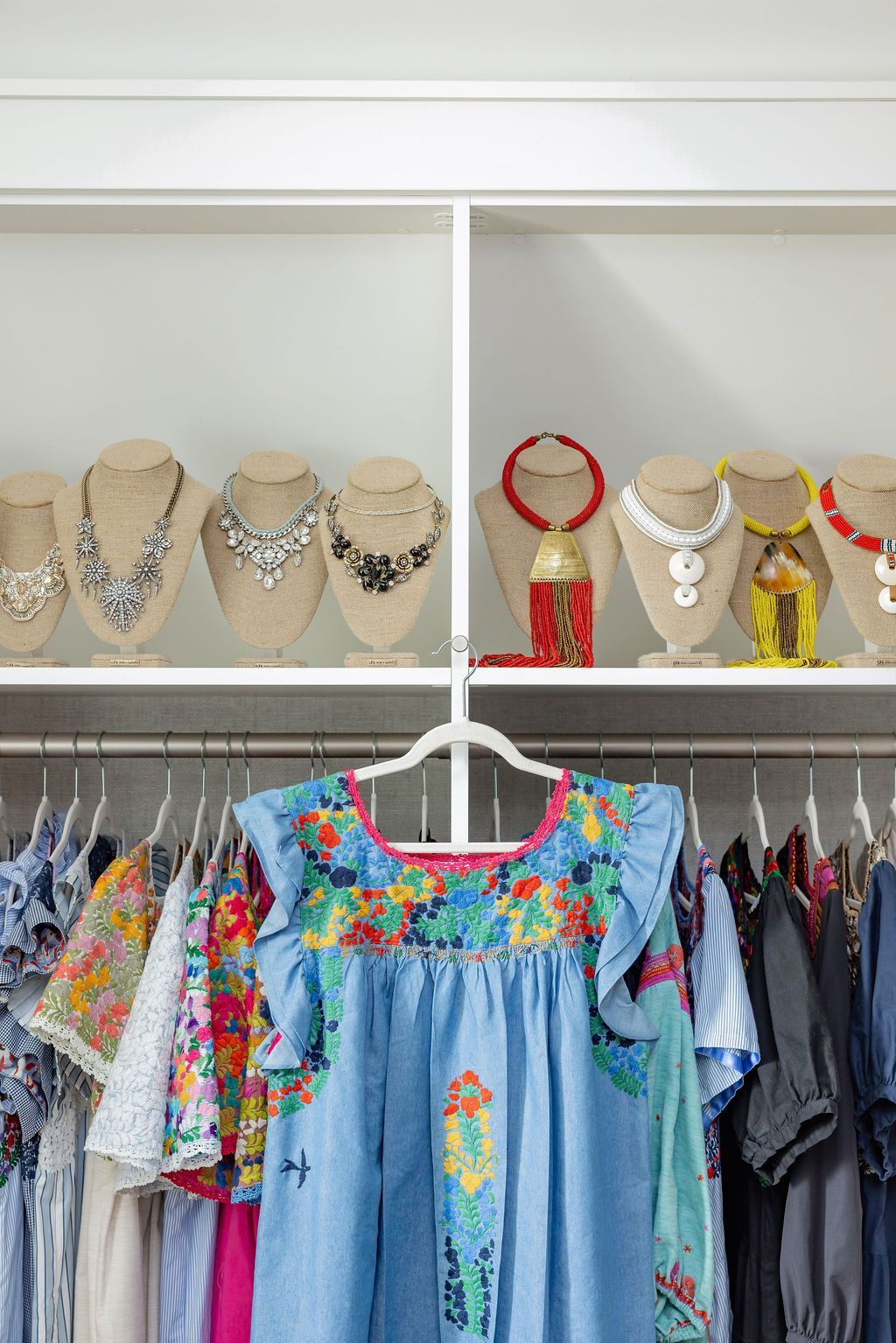 Closet with dresses and necklaces. A blue floral dress hangs on a rack. Jewelry is displayed on shelves.