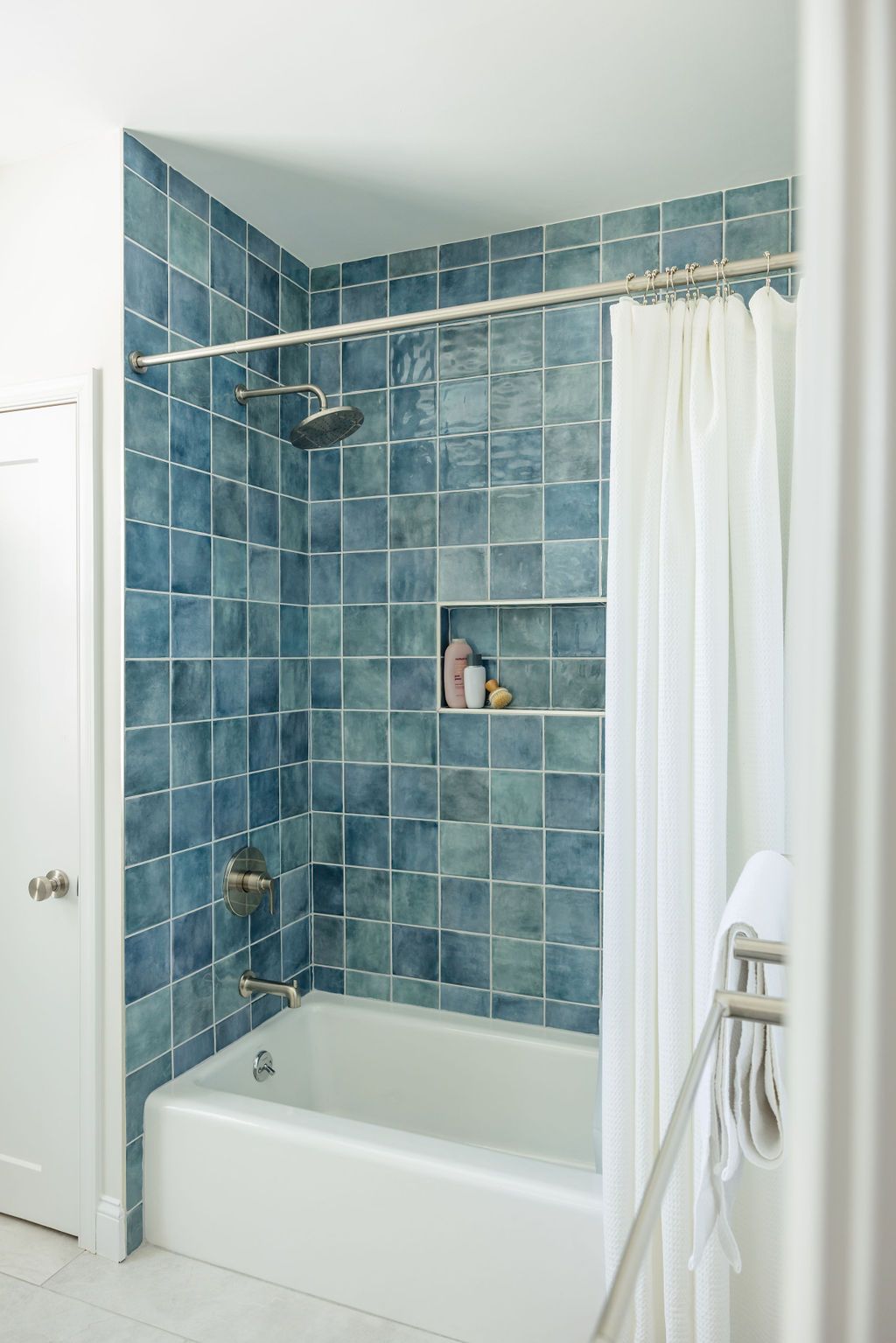 Blue-tiled shower with a white tub and curtain, recessed shelf with toiletries.