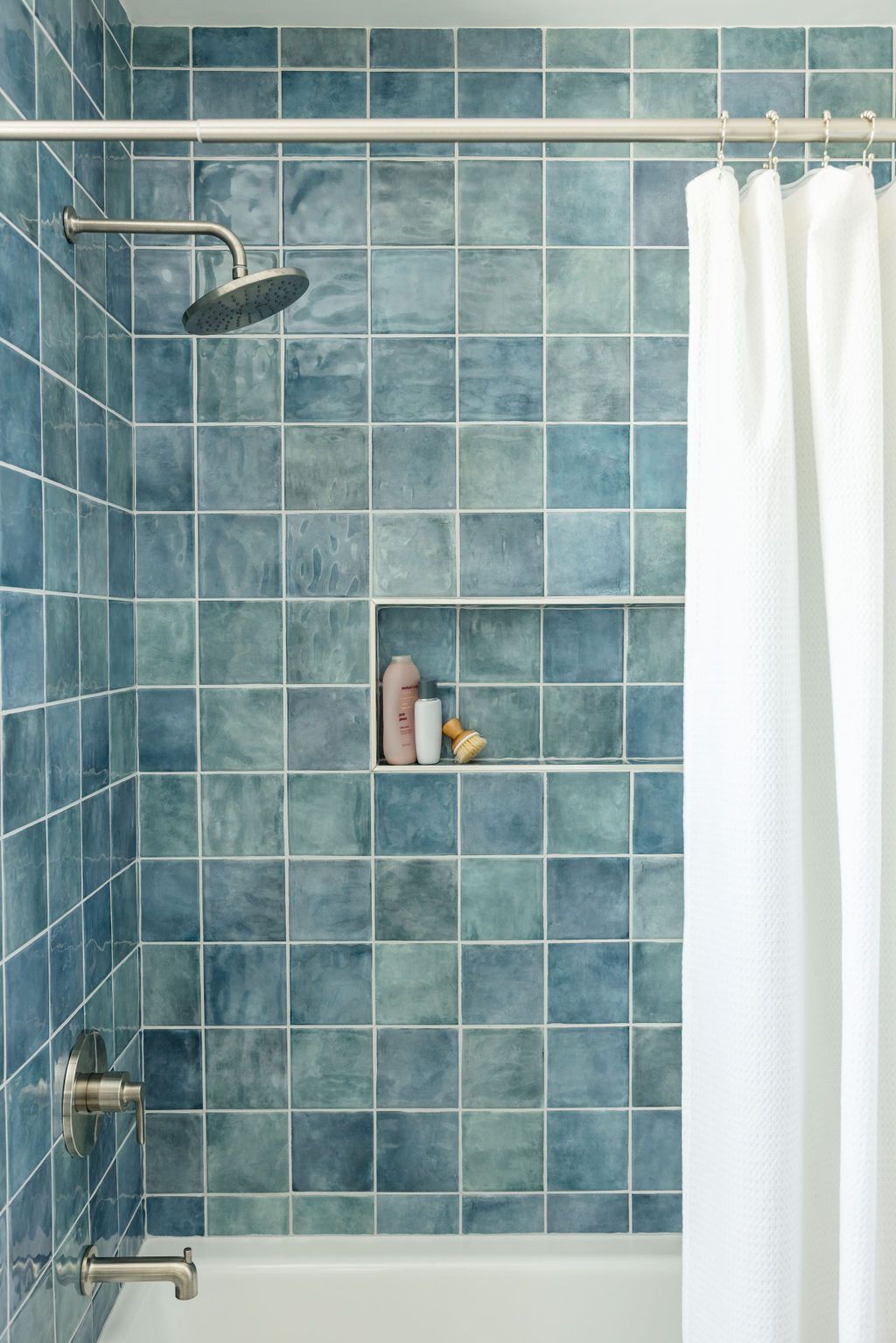 Blue tiled shower with white curtain, built-in shelf with toiletries, and a shower head.