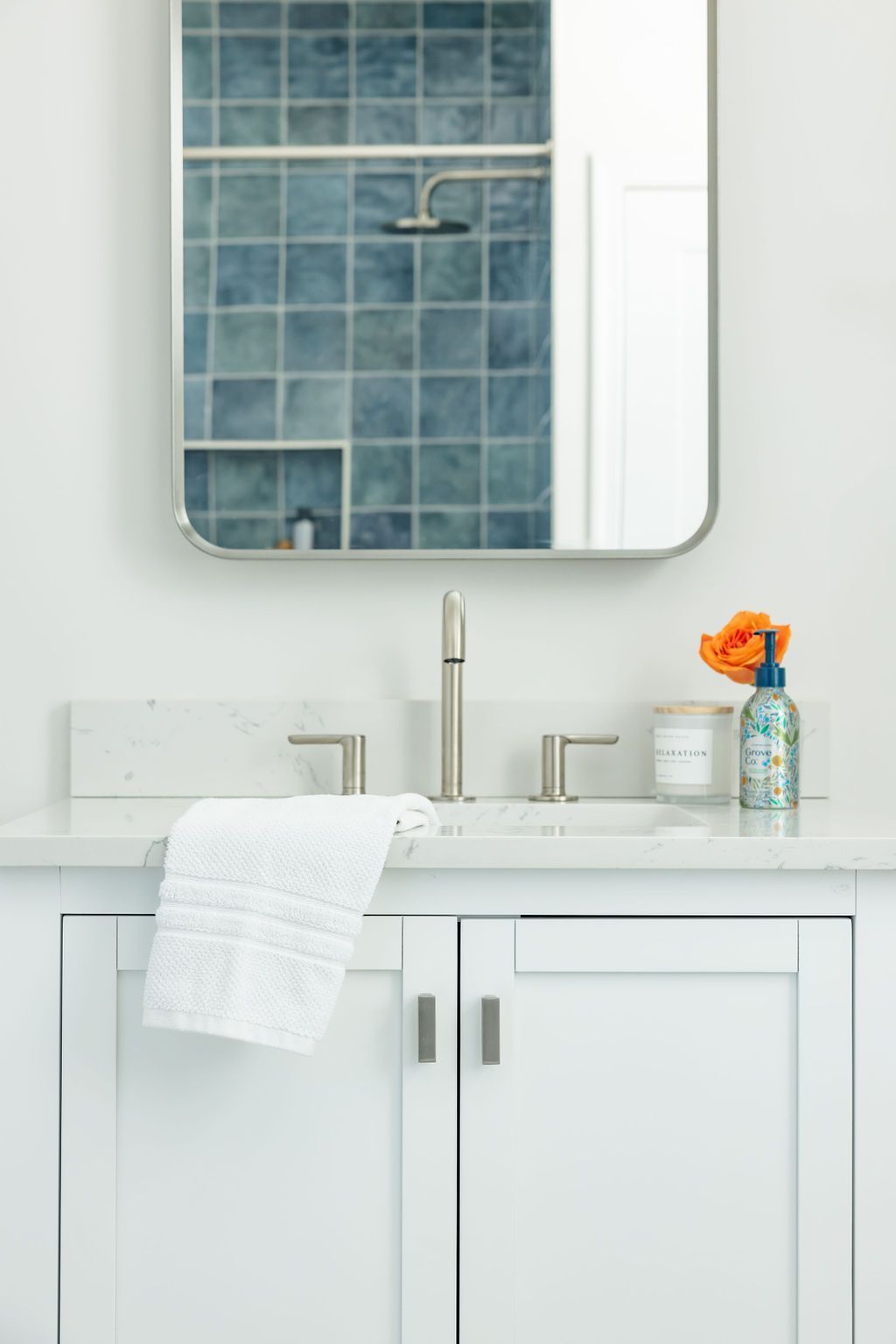 White bathroom vanity with silver fixtures, a white towel, and an orange flower. Blue tile shower reflected in the mirror.