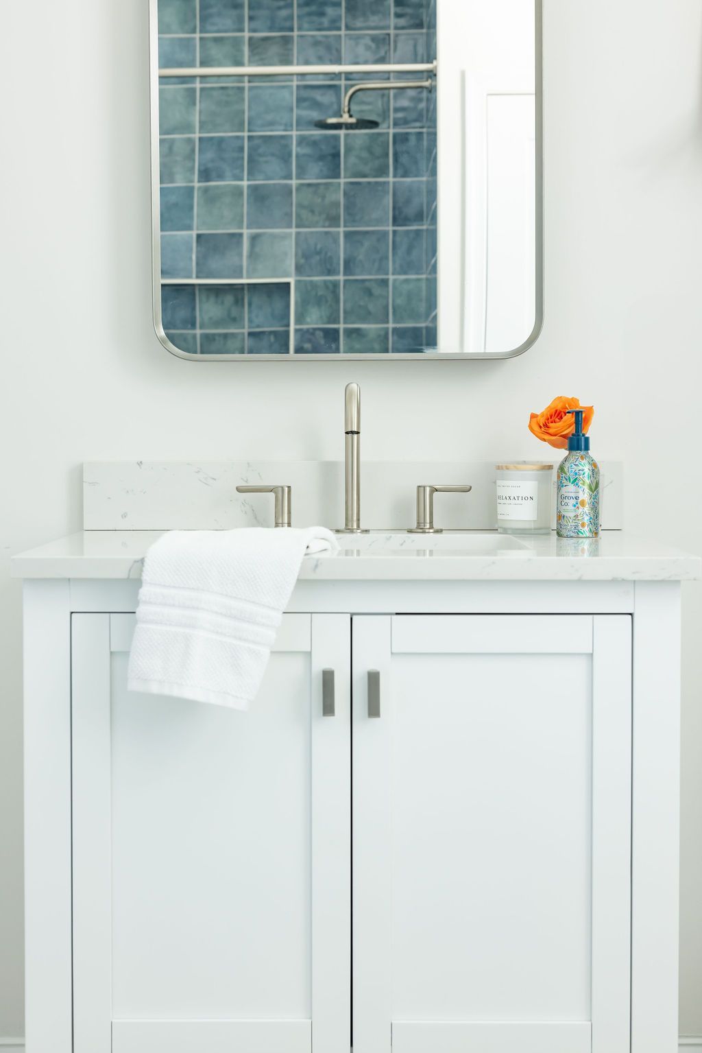 White bathroom vanity with sink, mirror, and toiletries. Blue tile shower visible in mirror.