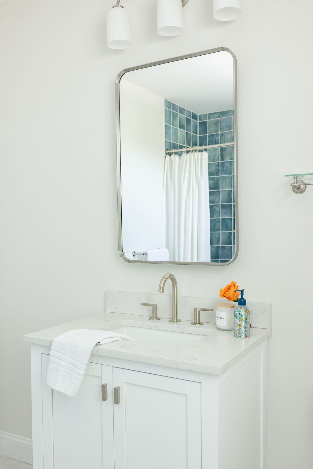White bathroom vanity with marble countertop, mirror, and faucet. White walls, blue tile shower visible in mirror.