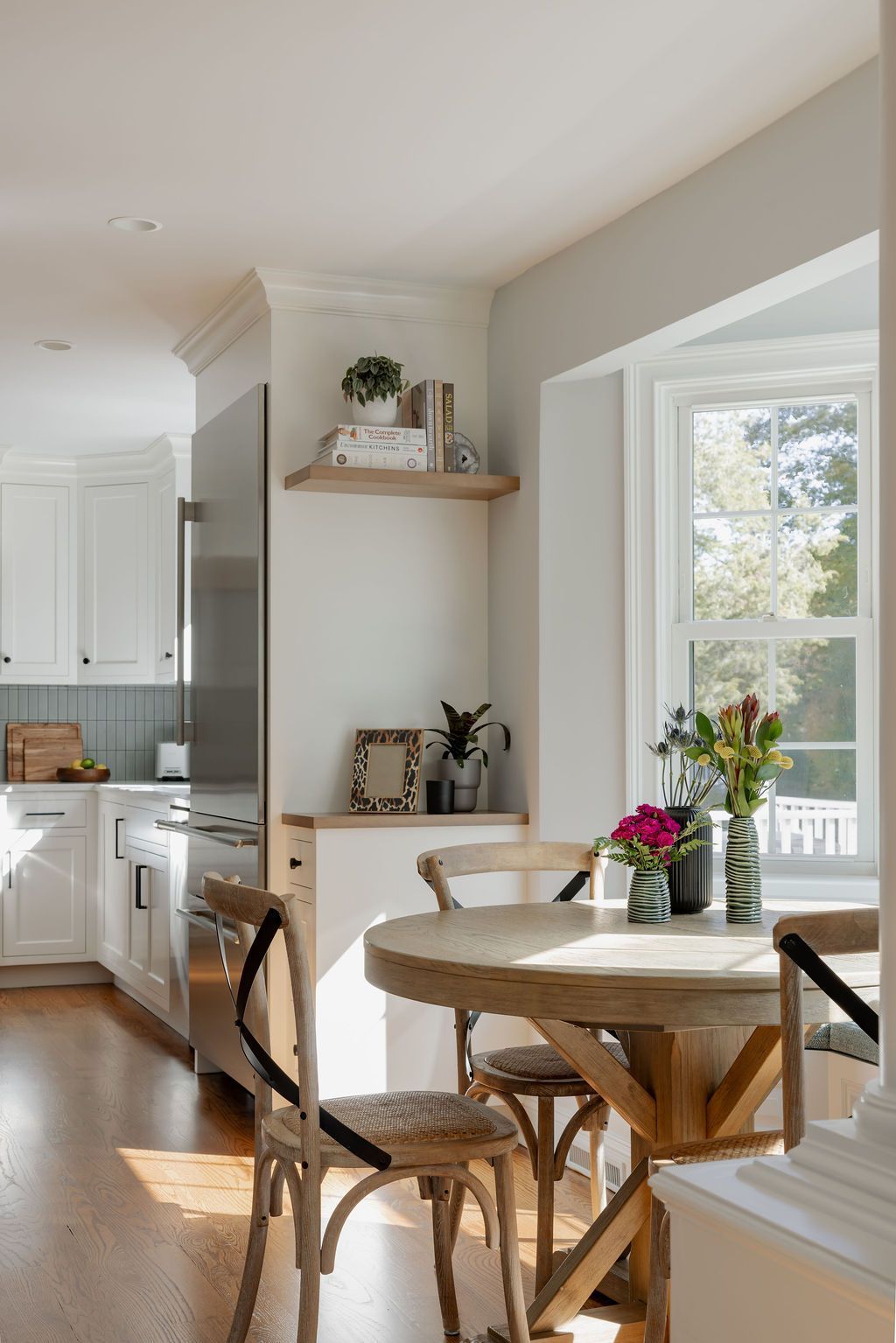 Bright kitchen with a round table and chairs near a window.  White cabinets and shelves, wood floor.