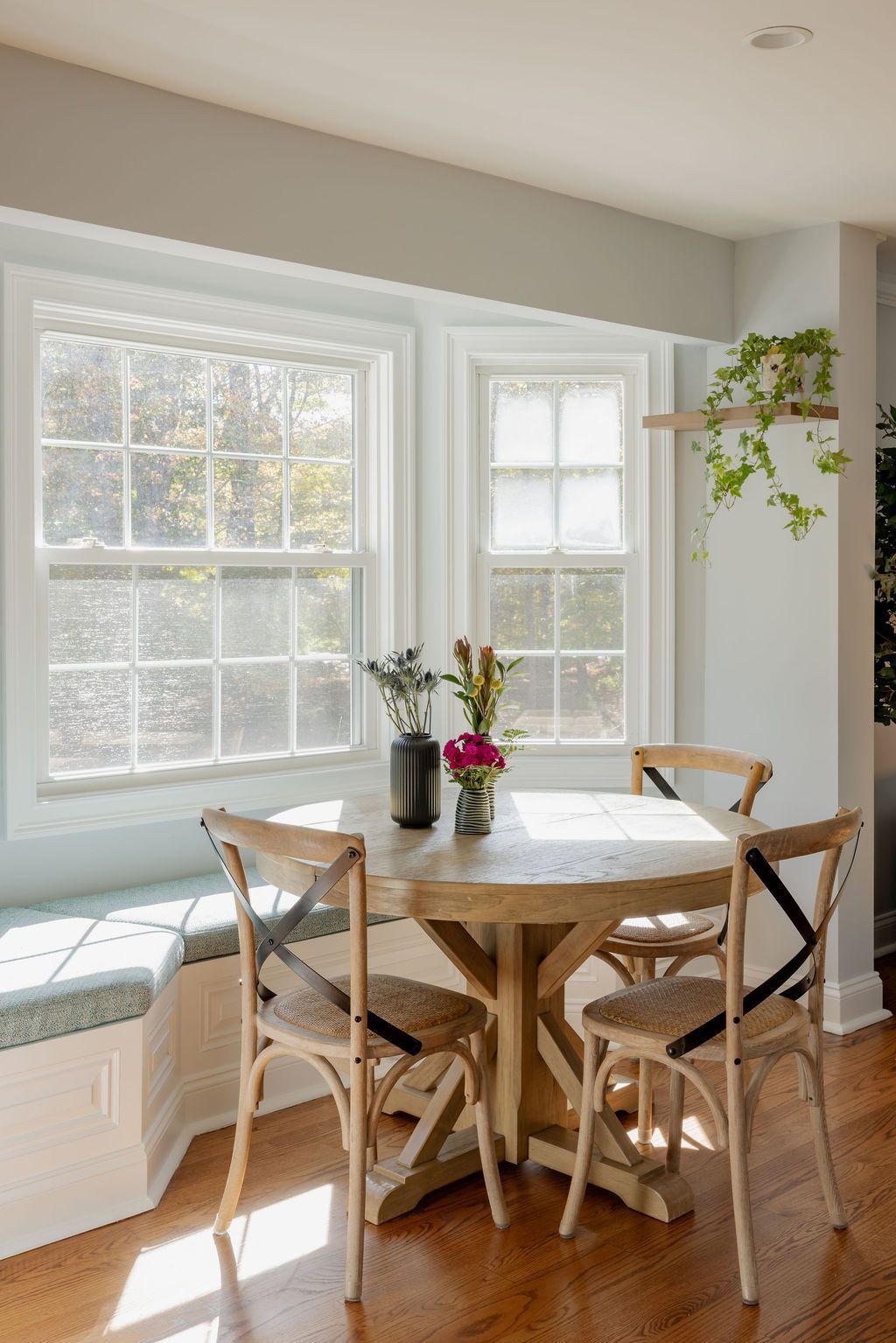 Dining area with a round wooden table, four chairs, and a window seat. Natural light fills the room.
