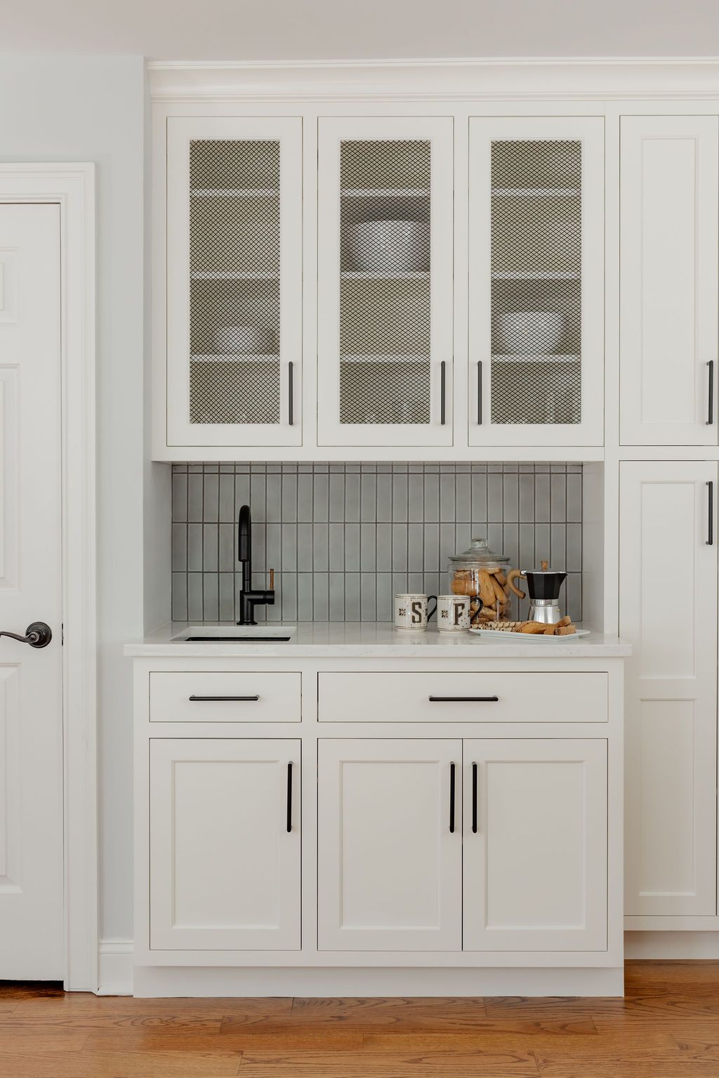 White built-in coffee station with upper glass-front cabinets and lower cabinets. Black faucet, backsplash, and hardware.