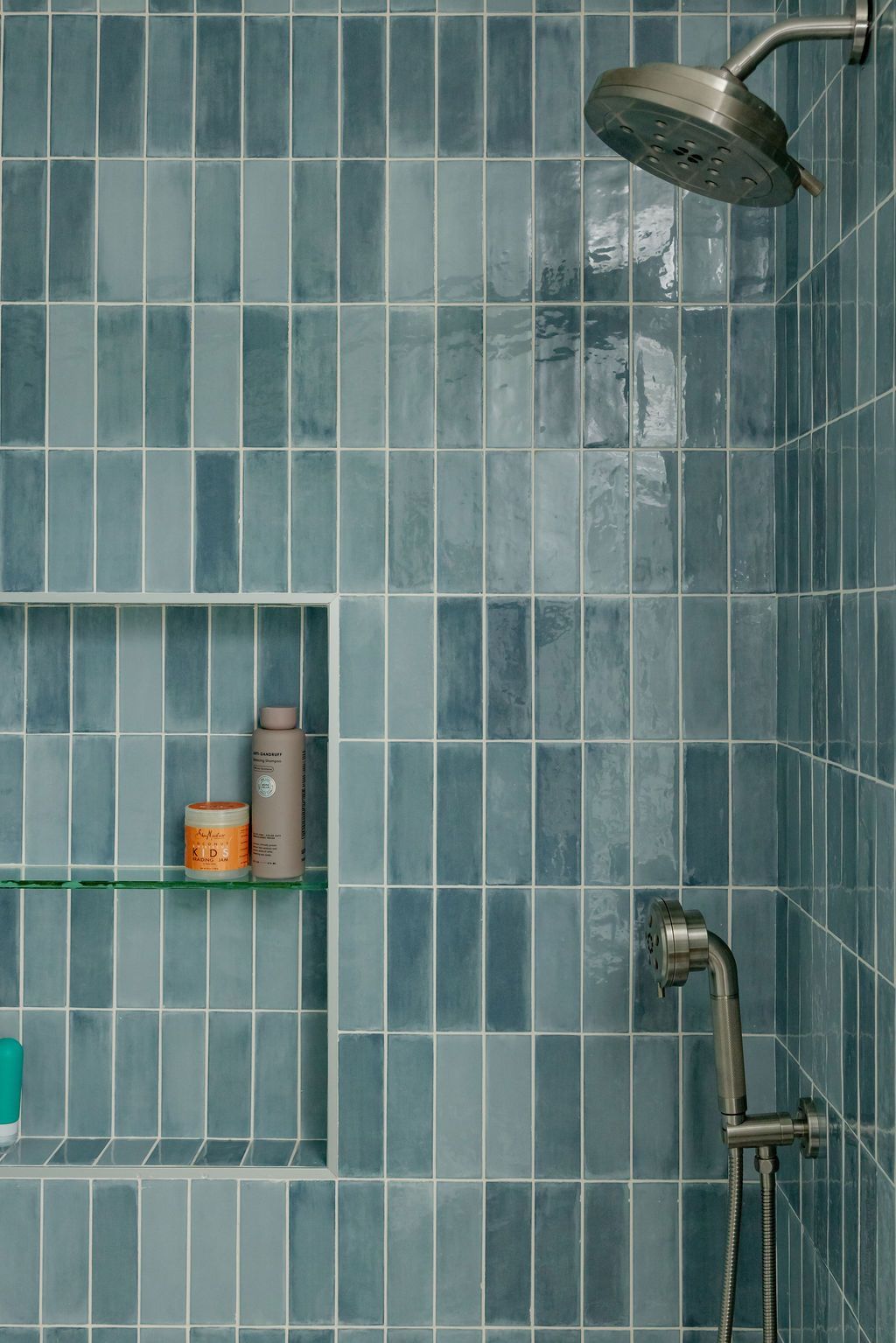 Blue tiled shower with a shelf, shower head, and handheld sprayer.