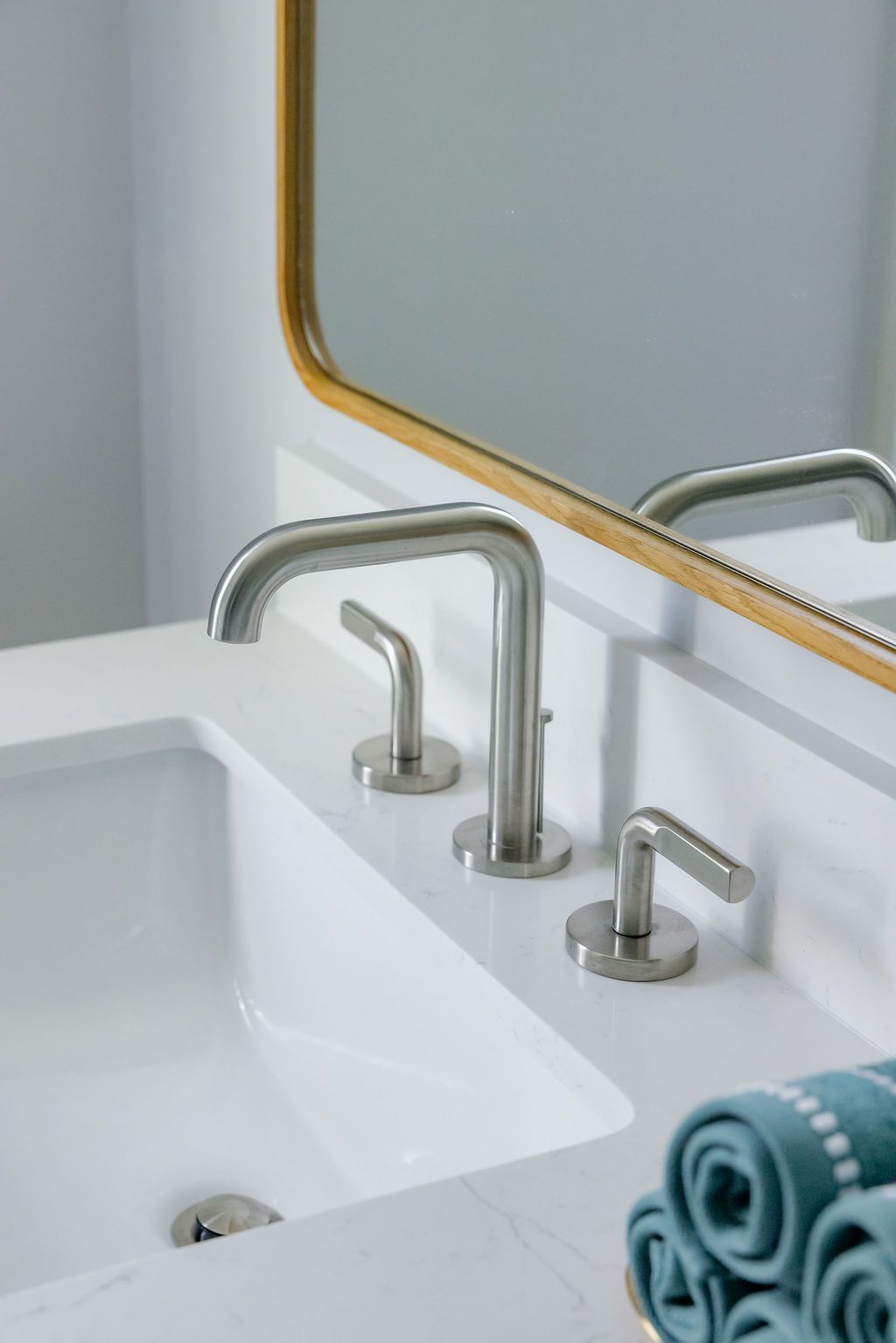 Bathroom sink with stainless steel faucet, white counter, and gold-framed mirror. Rolled towels in foreground.