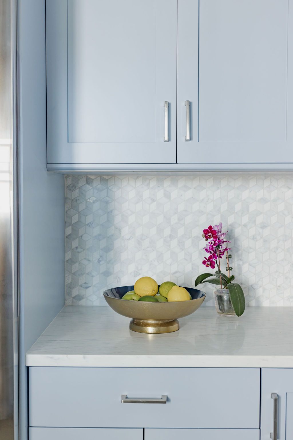 Kitchen countertop with fruit bowl and orchid, light blue cabinets, and patterned backsplash.