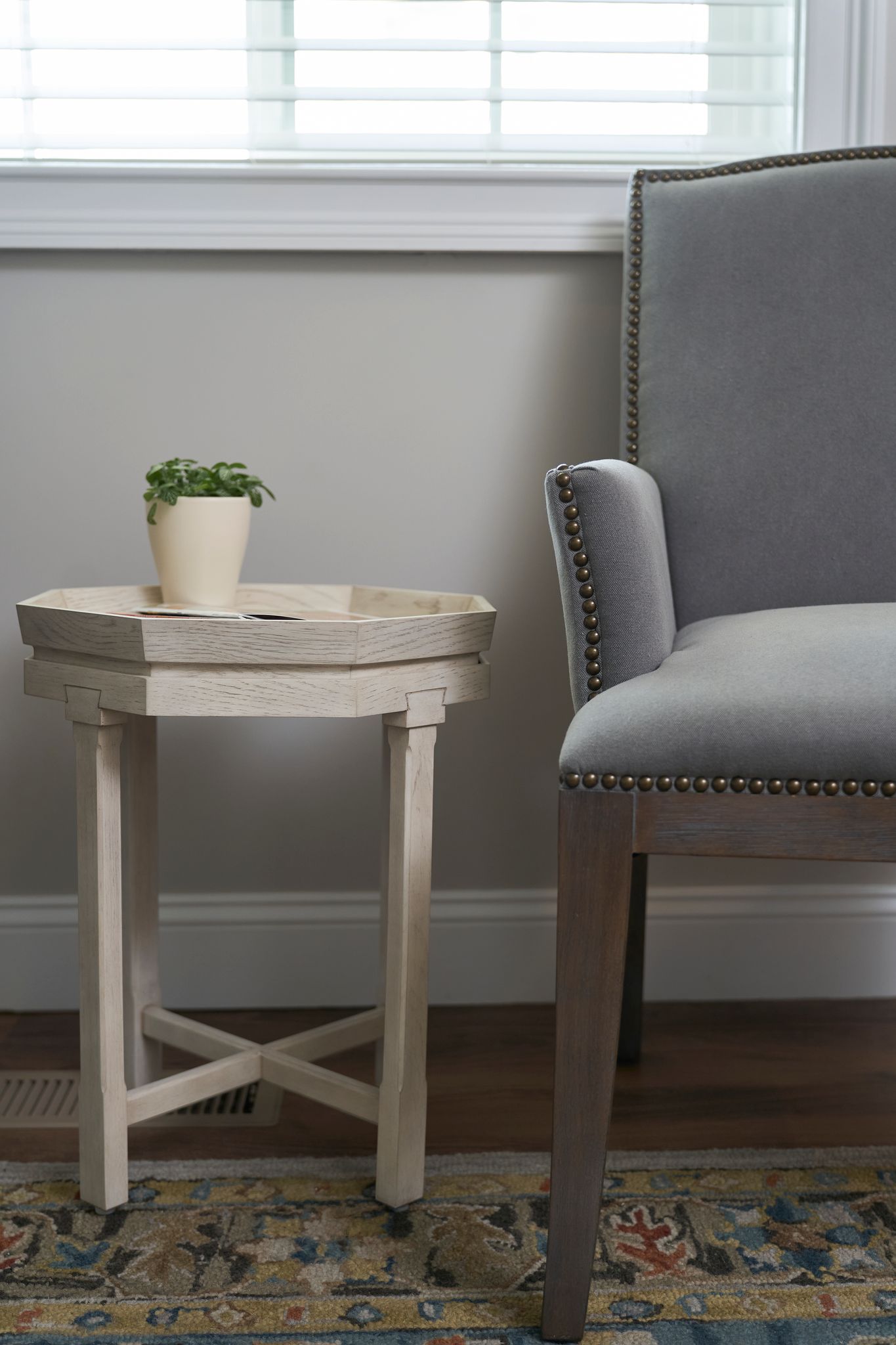 A small, white, octagonal side table with a potted plant next to a grey upholstered chair near a window.