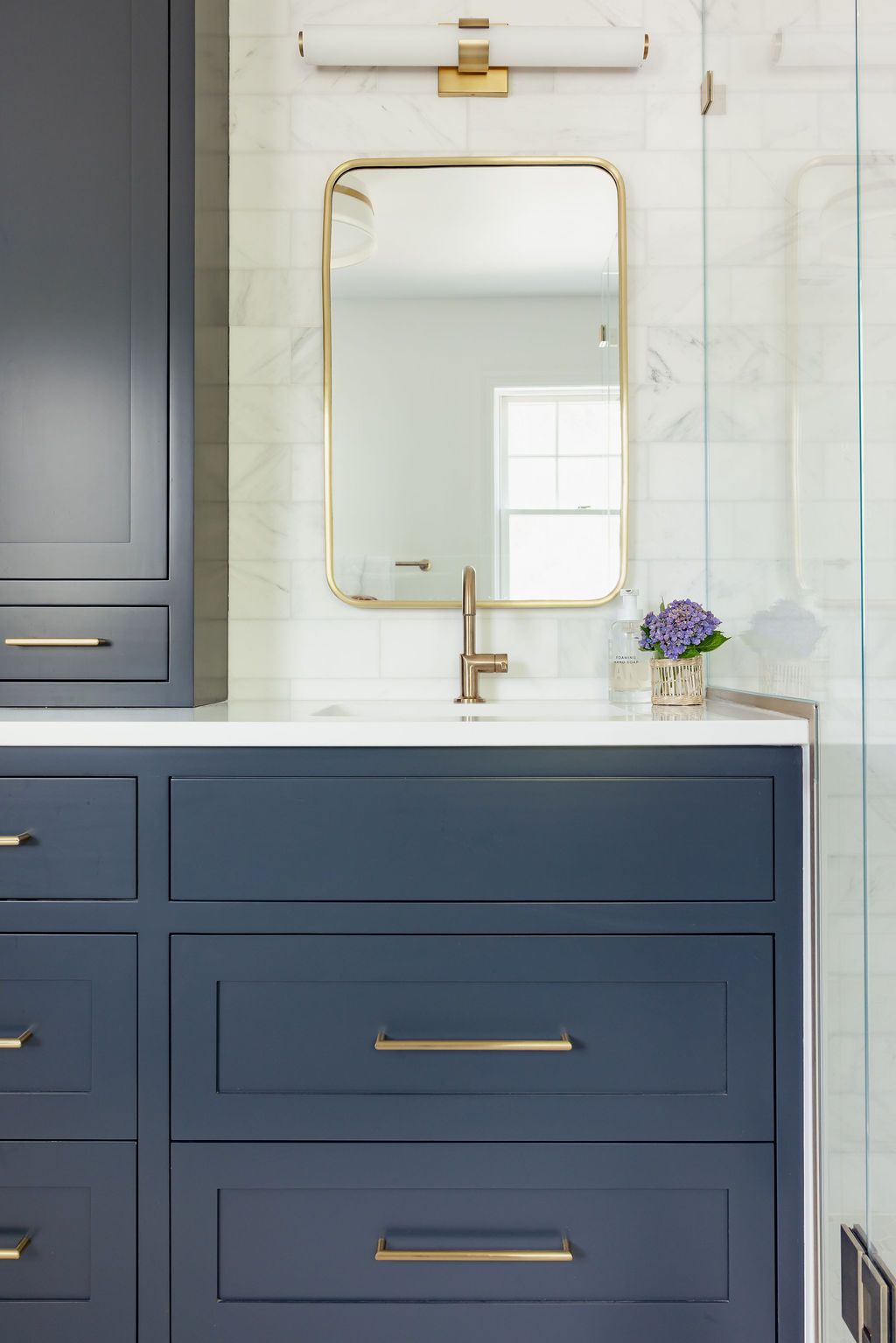 Blue vanity with gold hardware, marble backsplash, gold-framed mirror, and small purple flowers in bathroom.