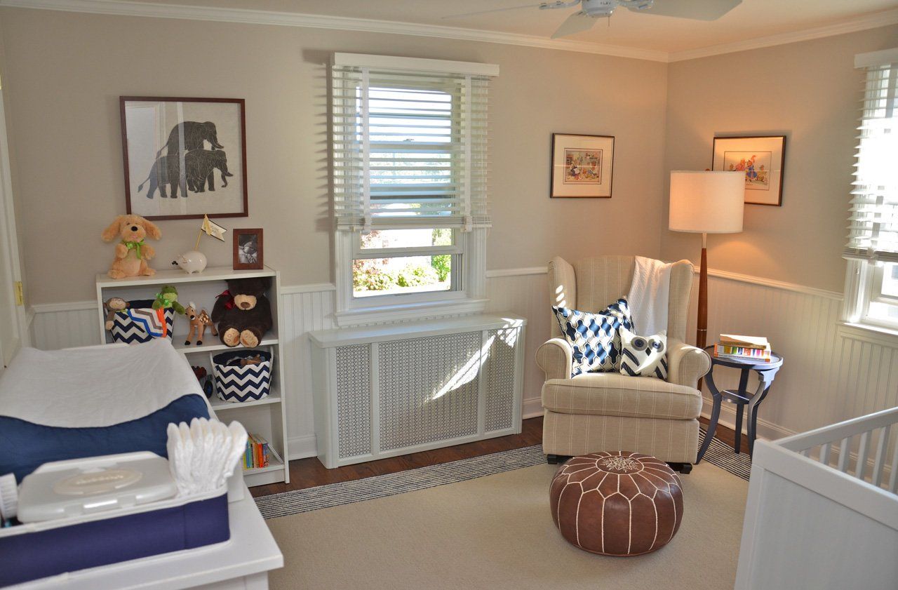 Nursery with neutral walls, cream armchair, dark blue accents, changing table, shelves, window, and a brown ottoman.