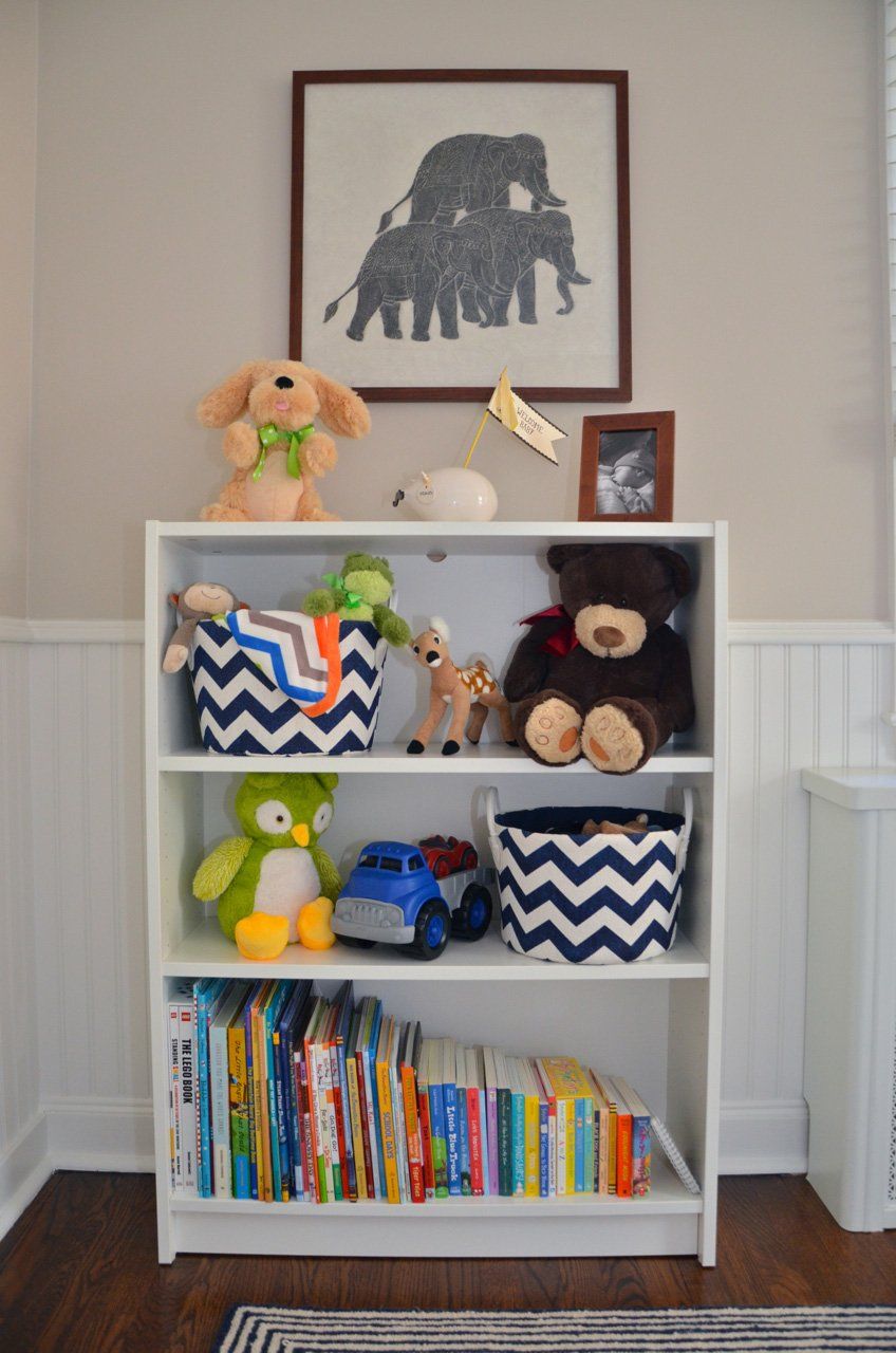 White bookshelf with toys and books, topped by an elephant art print, against a neutral wall.