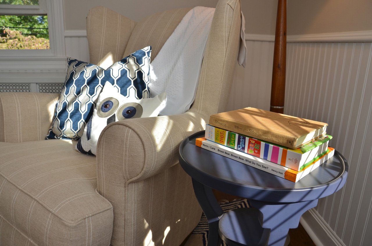 Cozy reading nook: beige armchair with patterned pillows, books on a blue side table, natural light from a window.