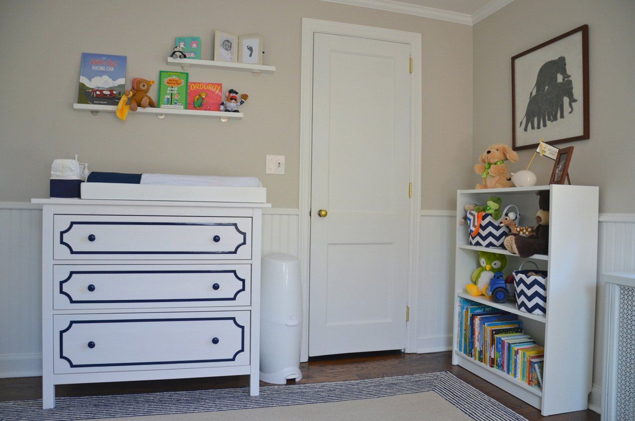 Nursery room with white dresser, changing table, and bookshelf. Books and toys are visible.