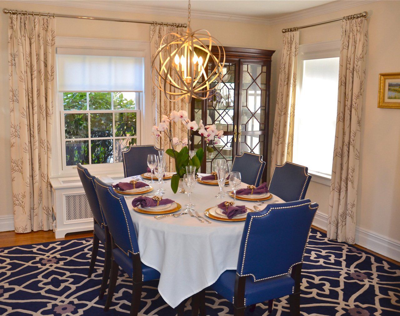 Dining room with round table set for eight, blue chairs, gold chandelier, and floral patterned curtains.